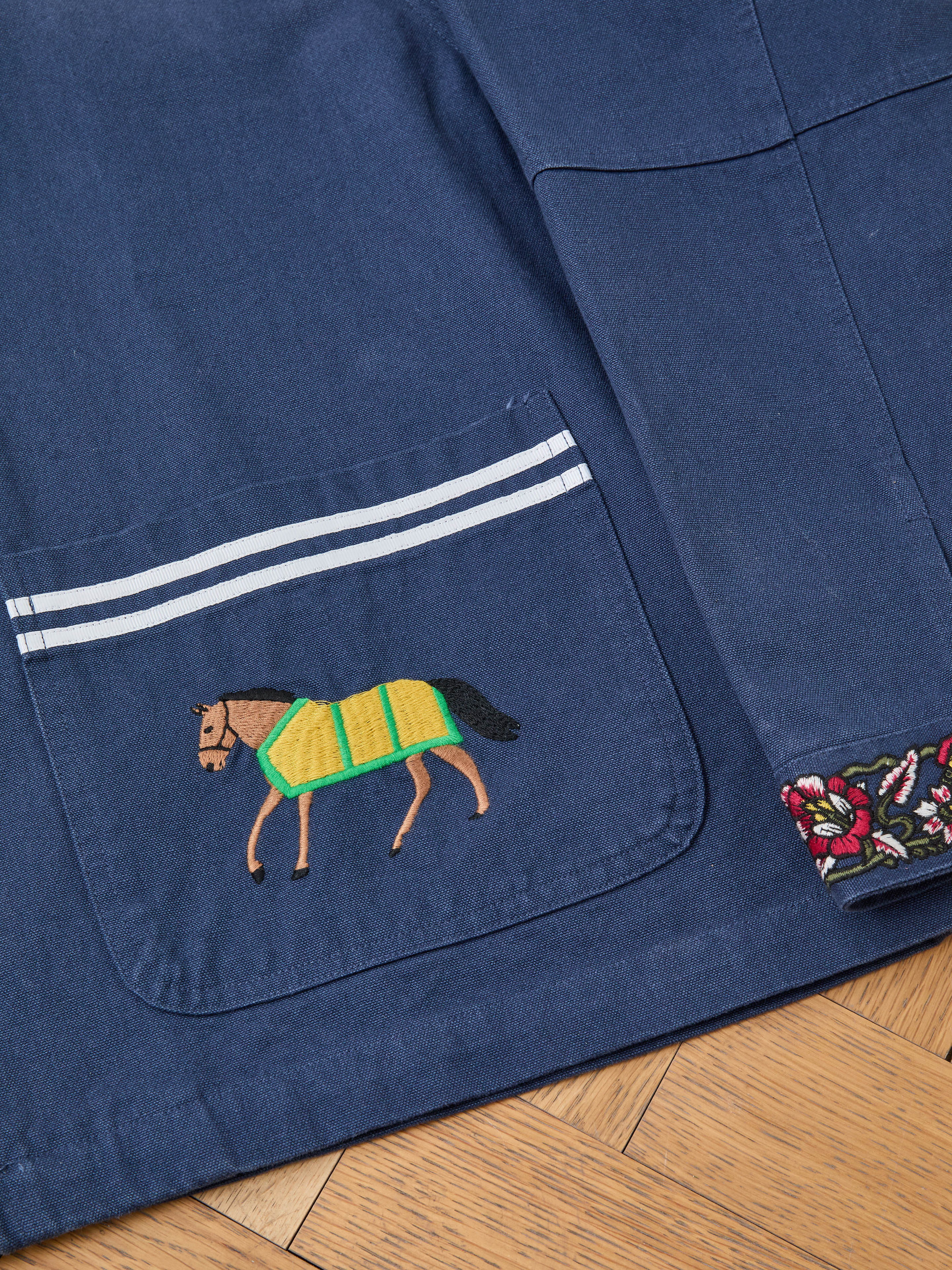 A close-up of the Found Horse Equine Work Jacket highlights its pocket with white stripes, an embroidered horse in racing colours with a yellow-green blanket, and a floral patterned cuff.