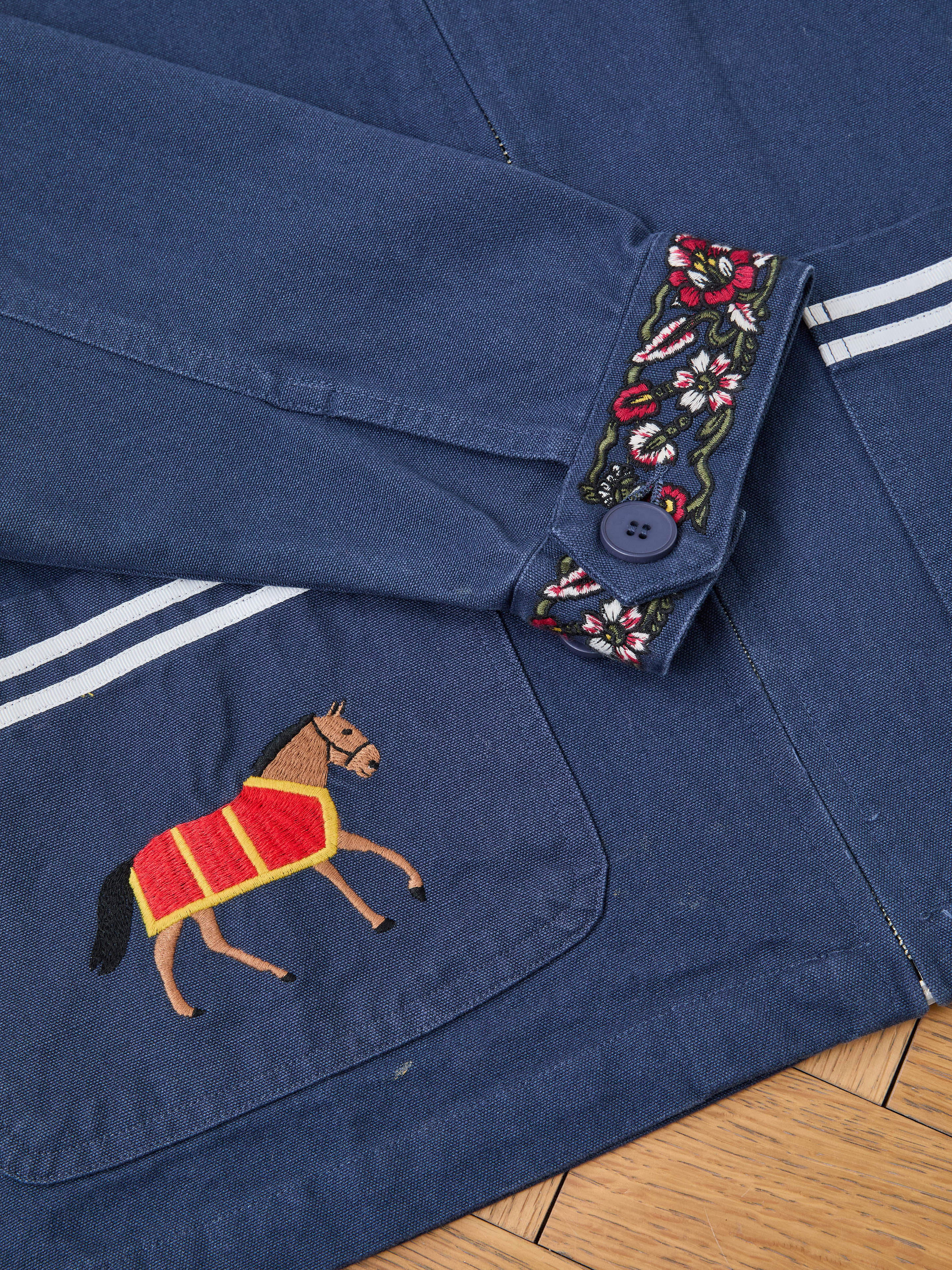 The Found Horse Equine Work Jacket is blue, with an embroidered horse on the pocket, floral embroidery on the cuff, and a button detail.