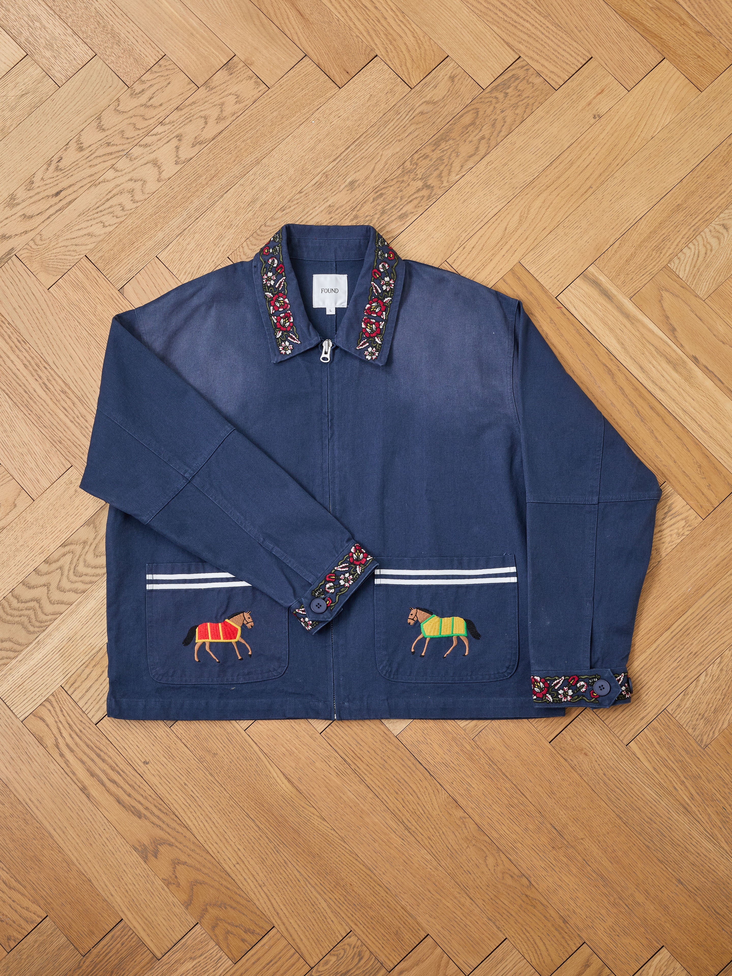 The Found Horse Equine Work Jacket, featuring embroidered horses on the front pockets and decorative patterns on the collar and cuffs, is showcased on a wooden herringbone floor.