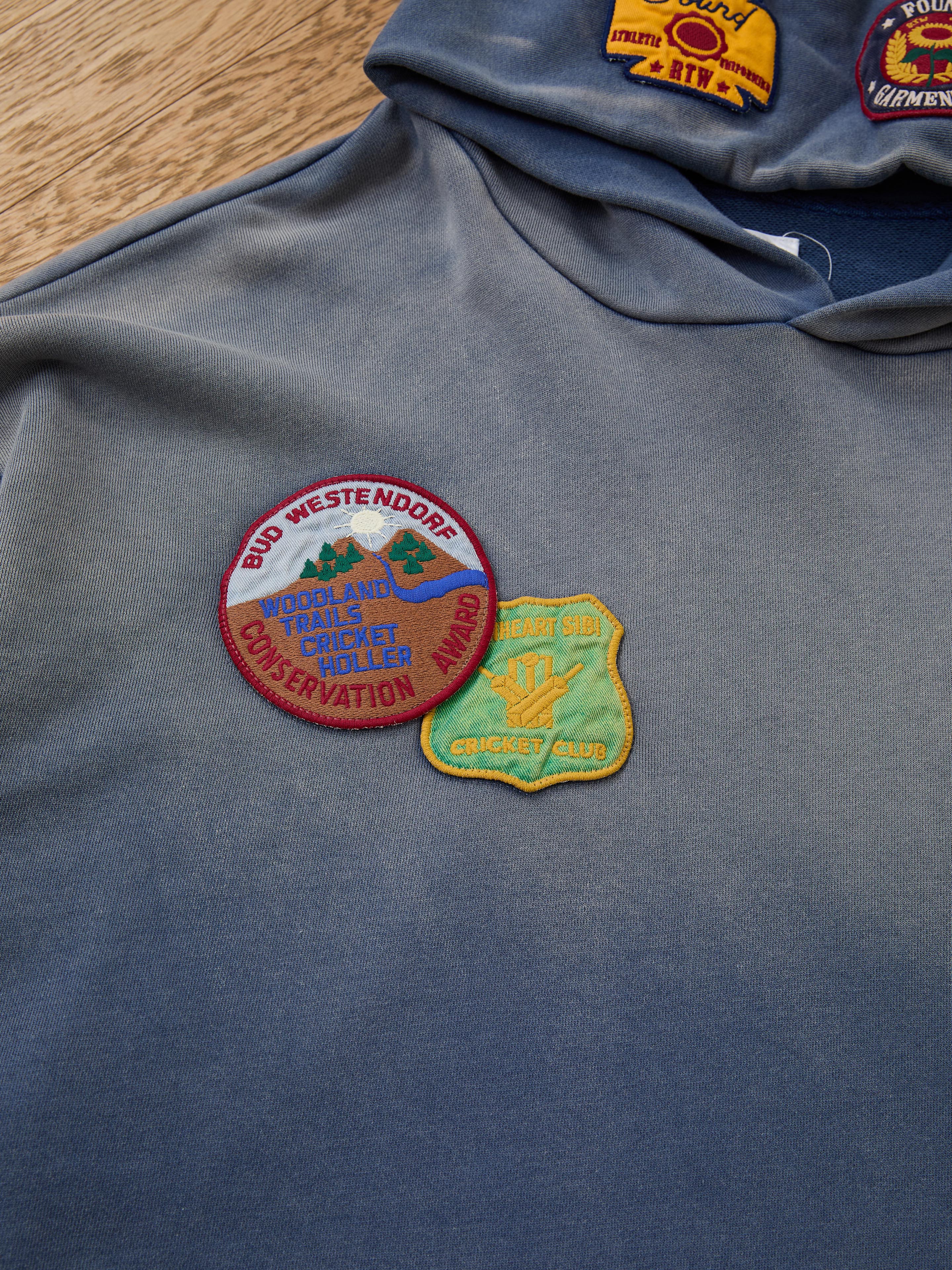The FOUND Distressed Patch Hoodie in blue is crafted from soft French Terry cotton, featuring two embroidered patches: a circular "Woodland Trails Conservation Award" patch and a yellow "Heart Gibi Cricket Club" badge.