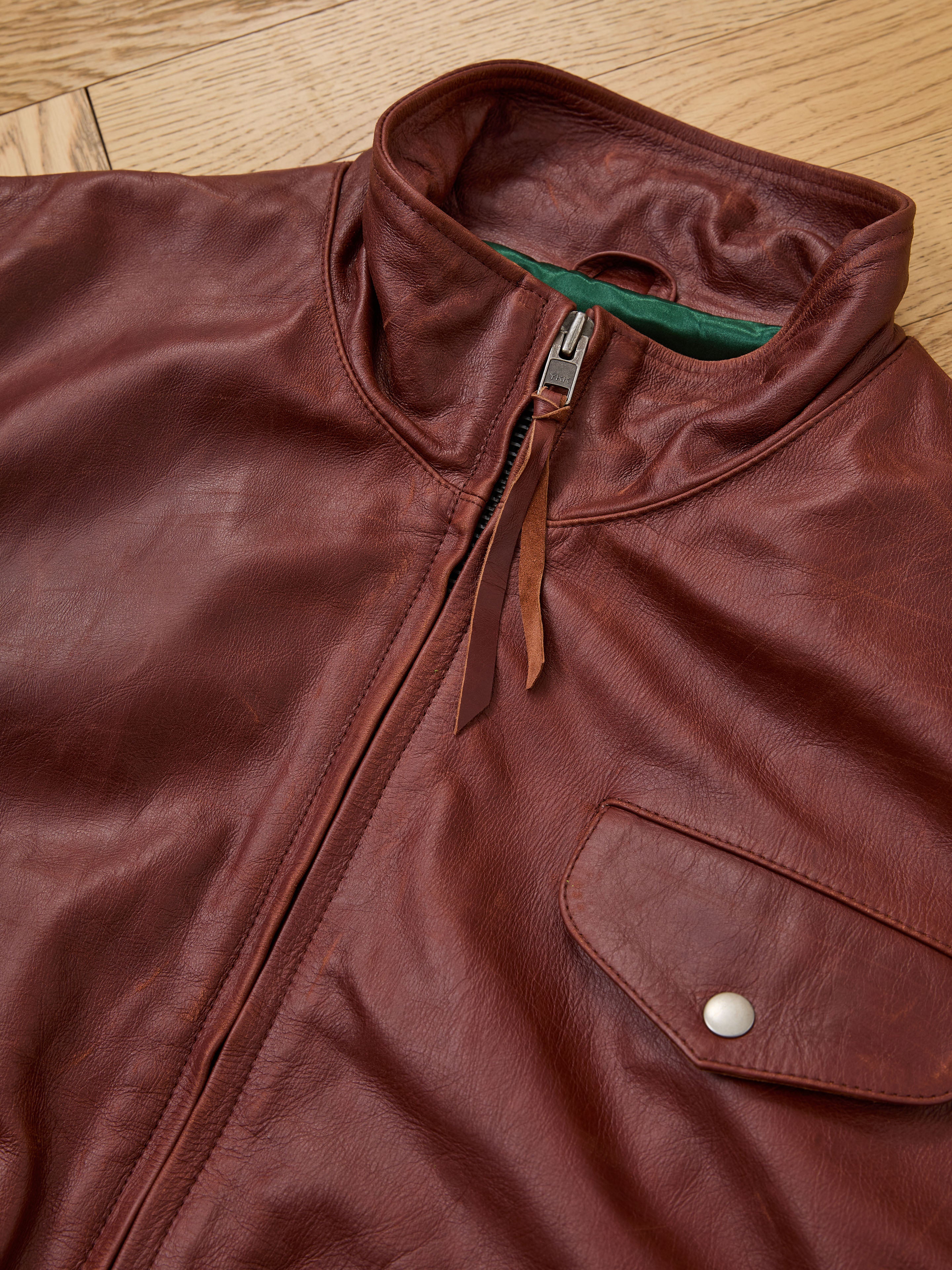 The Found Redwood Leather Bomber Jacket, featuring a front zipper, leather pull tabs, high collar, and buttoned chest pocket, is shown in a close-up on a wooden surface.