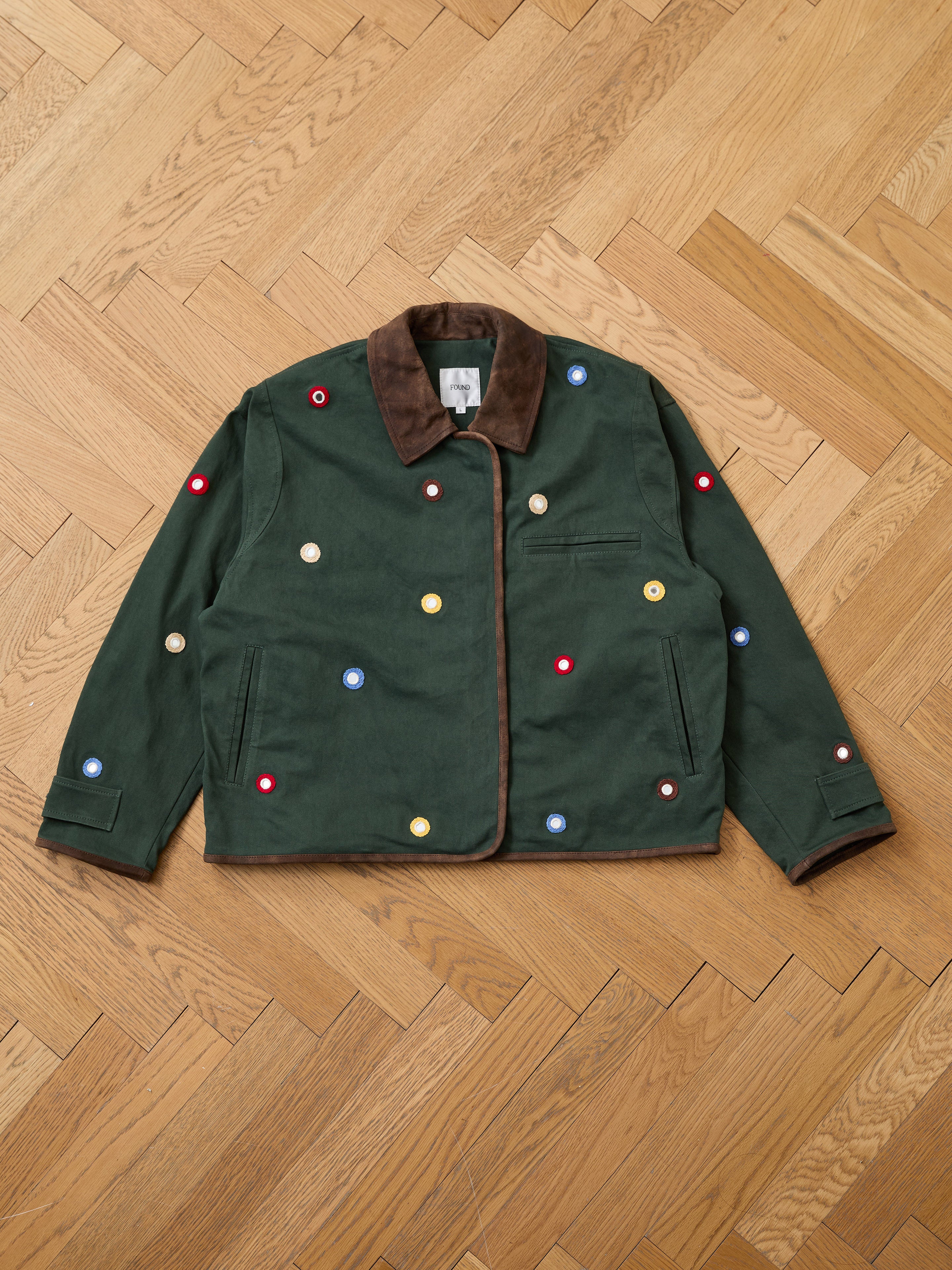 The Hand Embroidered Mirror Abhala Jacket by Found, a green artisan-made piece with a brown collar and multicolored circular patches, is displayed flat on a herringbone-patterned wooden floor.