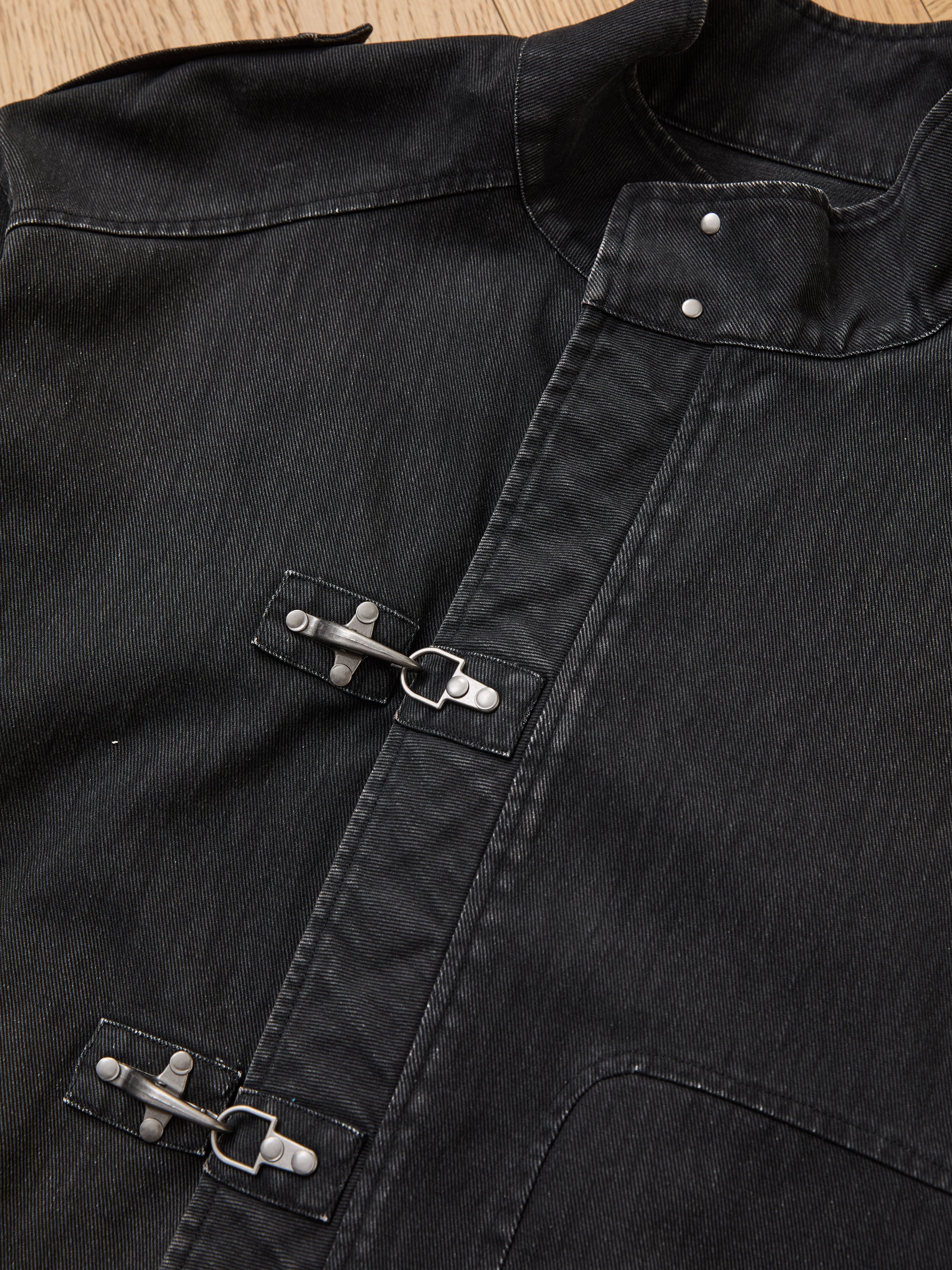 A close-up of the Found Utility Clasp Work Jacket, crafted from cotton twill with two silver clasp fasteners and metal rivets, laid on a wooden surface.