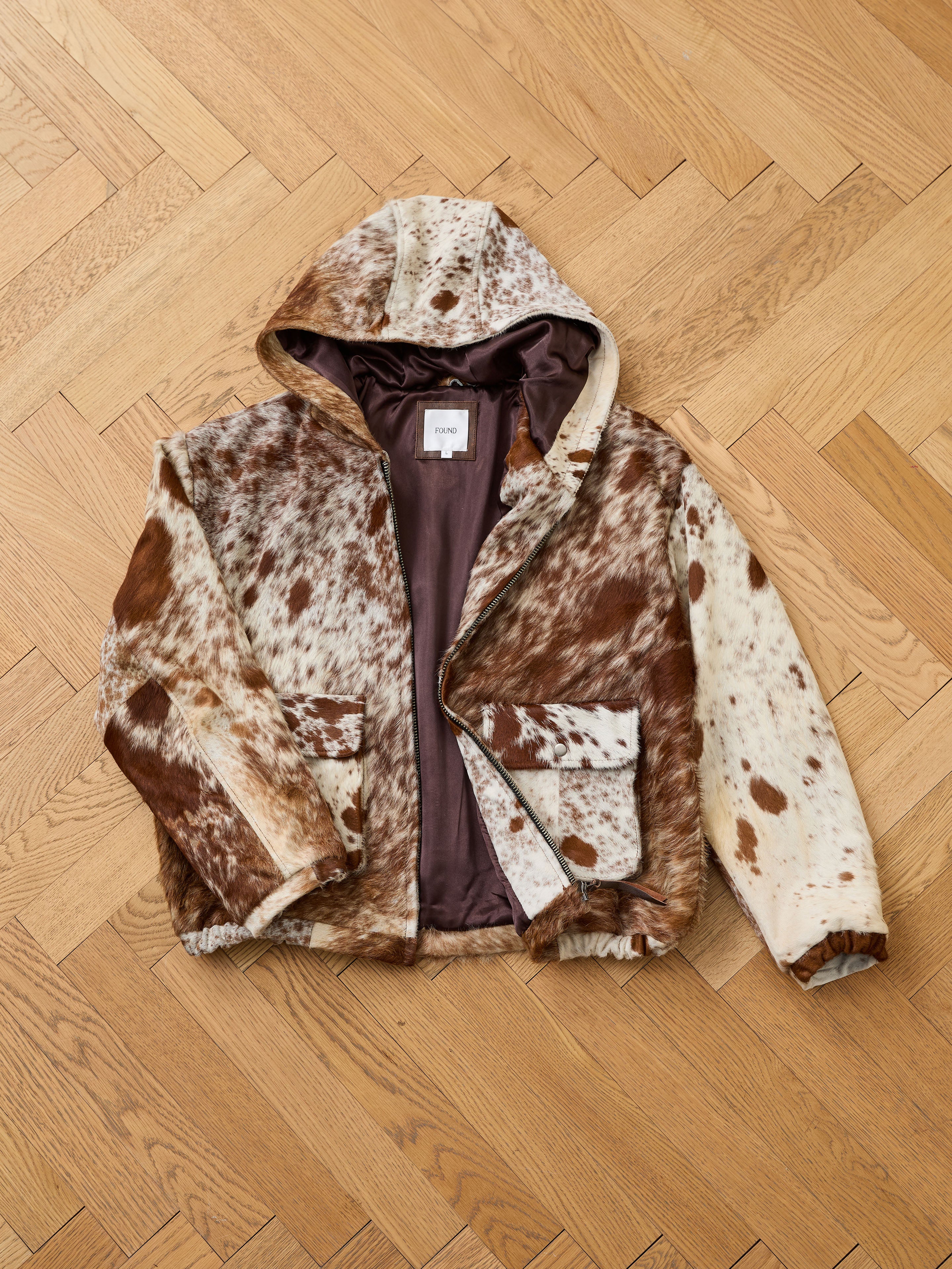 The FOUND Cowhide Drover Hoodie features a brown and white faux fur spotted pattern, front pockets, and visible lining, shown laid flat on a wooden herringbone floor.