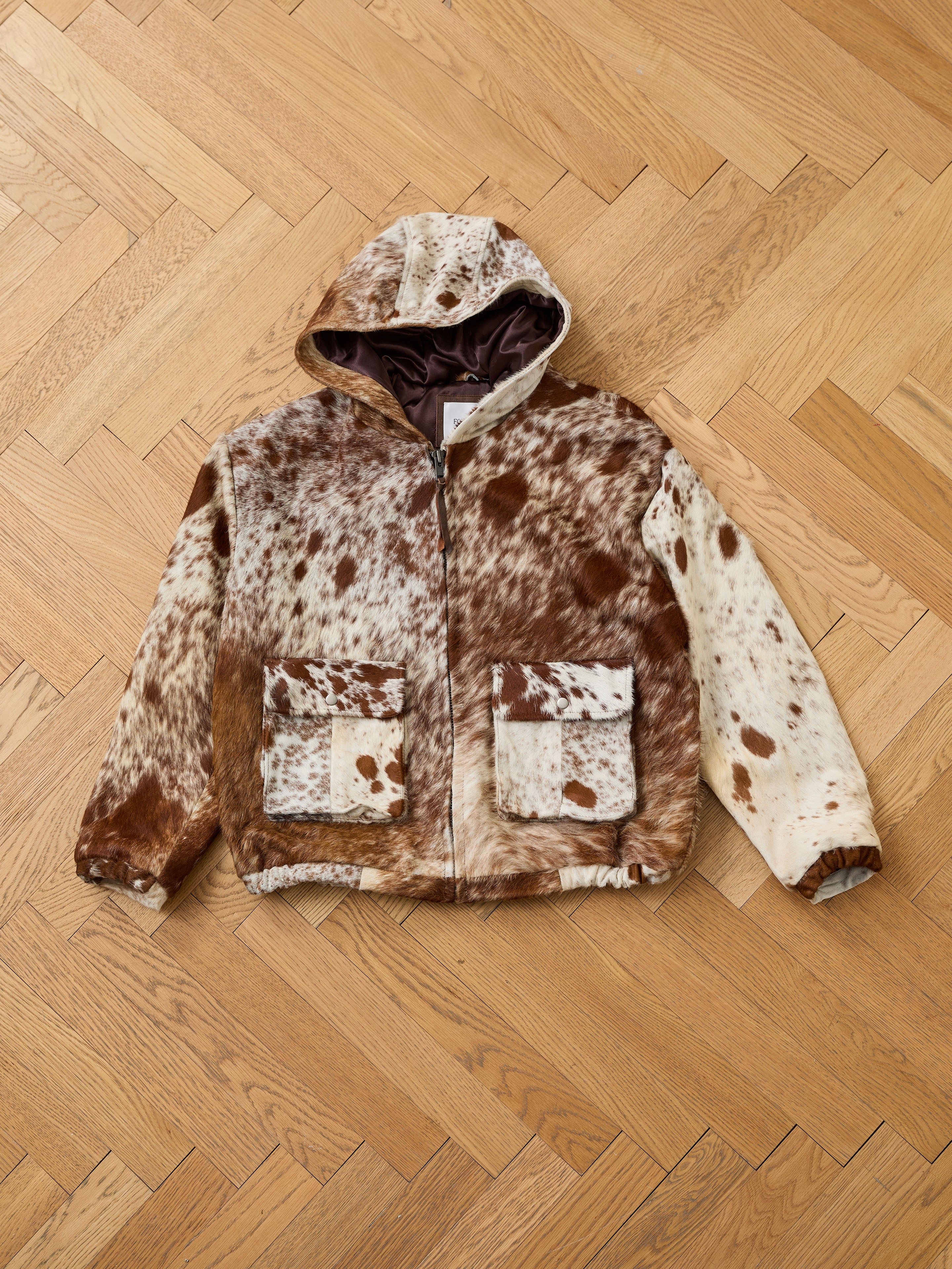 The FOUND Cowhide Drover Hoodie features a brown and white cow print, hooded zip-up design, and two front pockets, displayed on a light wood herringbone floor.