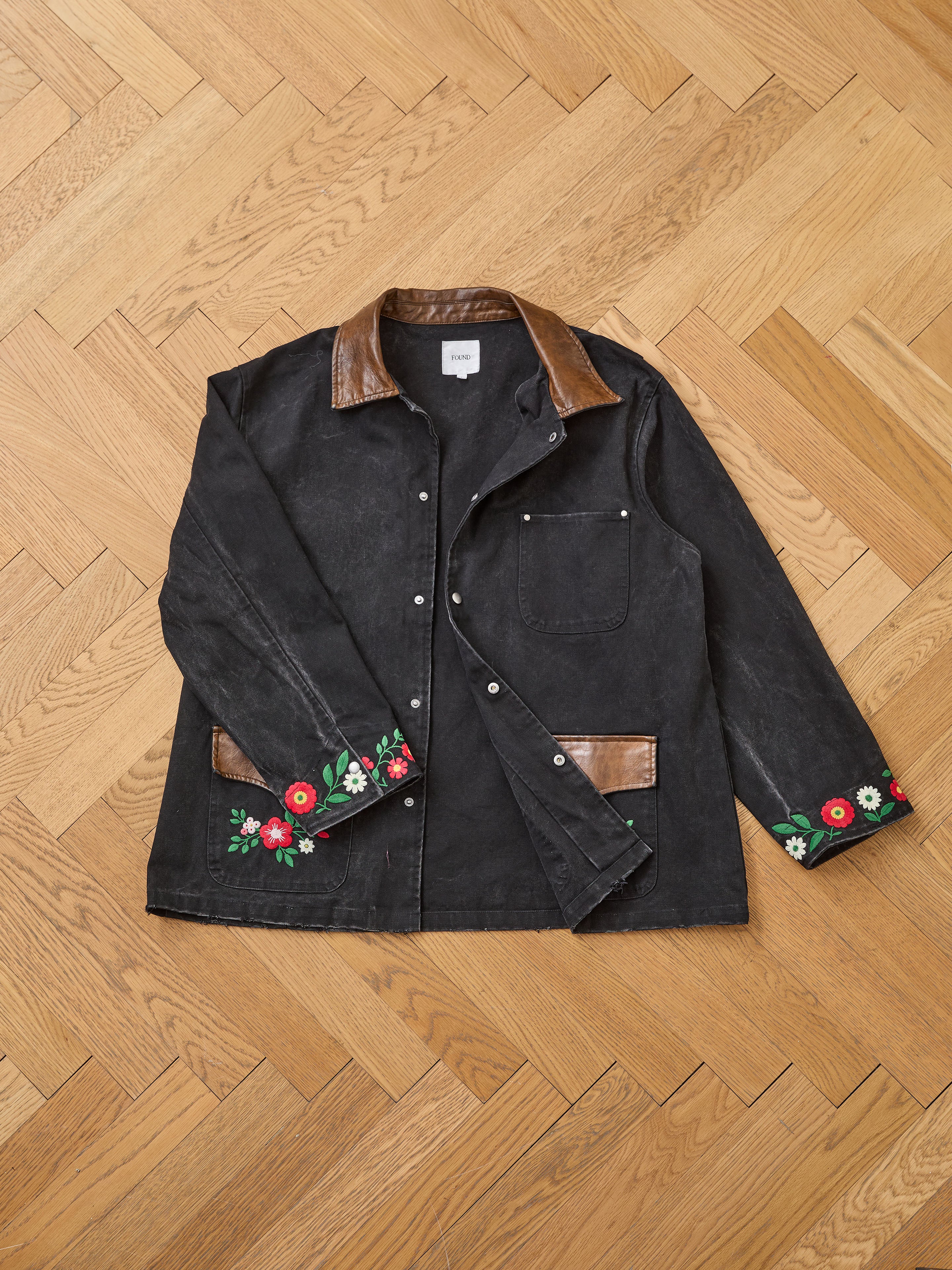 The Found Canvas Embroidered Work Jacket, featuring black canvas, brown leather trim on collar and cuffs, snap buttons, a front pocket, and floral sleeve embroidery, is laid flat on a wooden parquet floor.