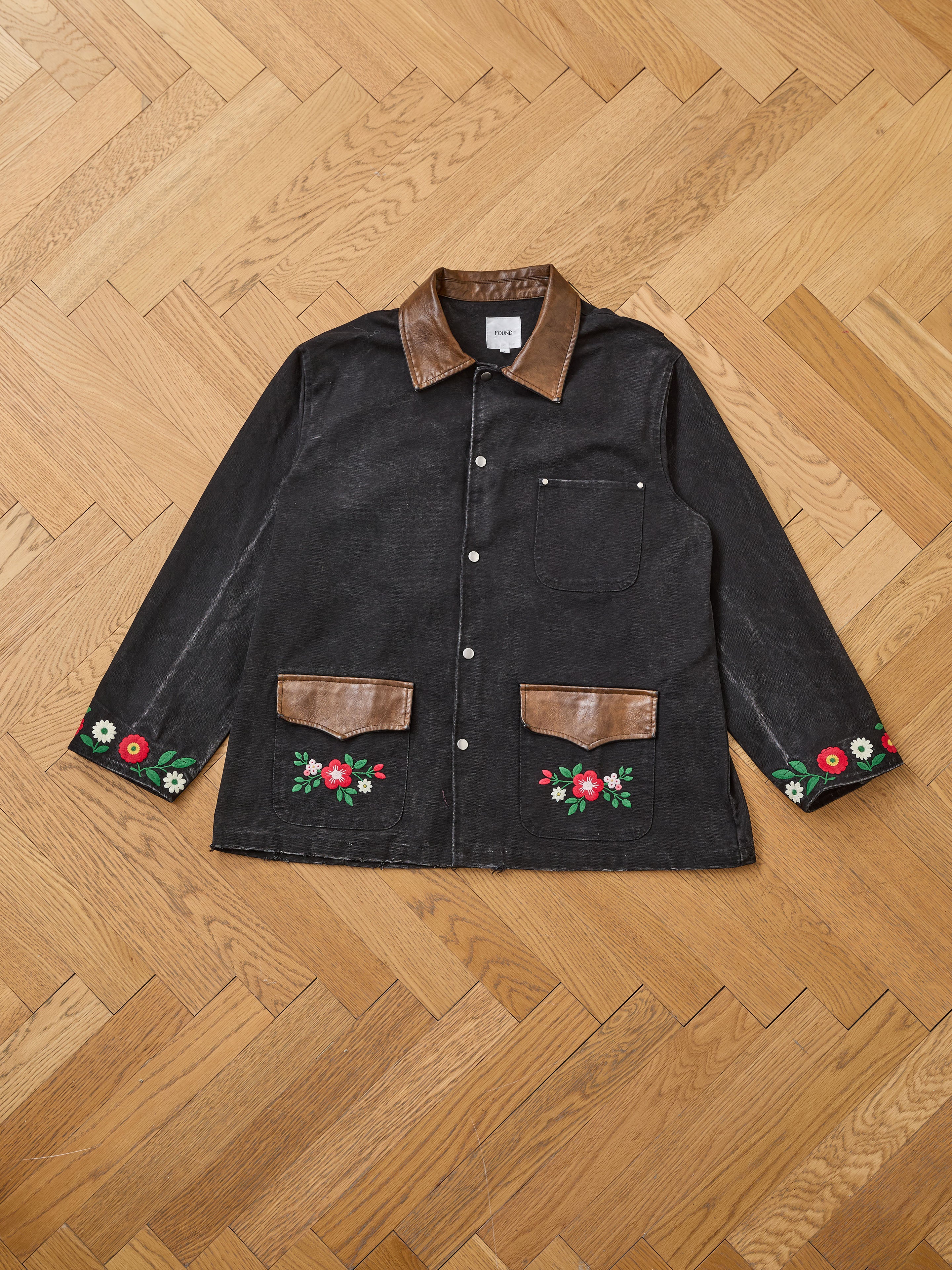 The Found Canvas Embroidered Work Jacket in black features brown leather trim and floral embroidery on the sleeves and pockets, shown on a wooden parquet floor.