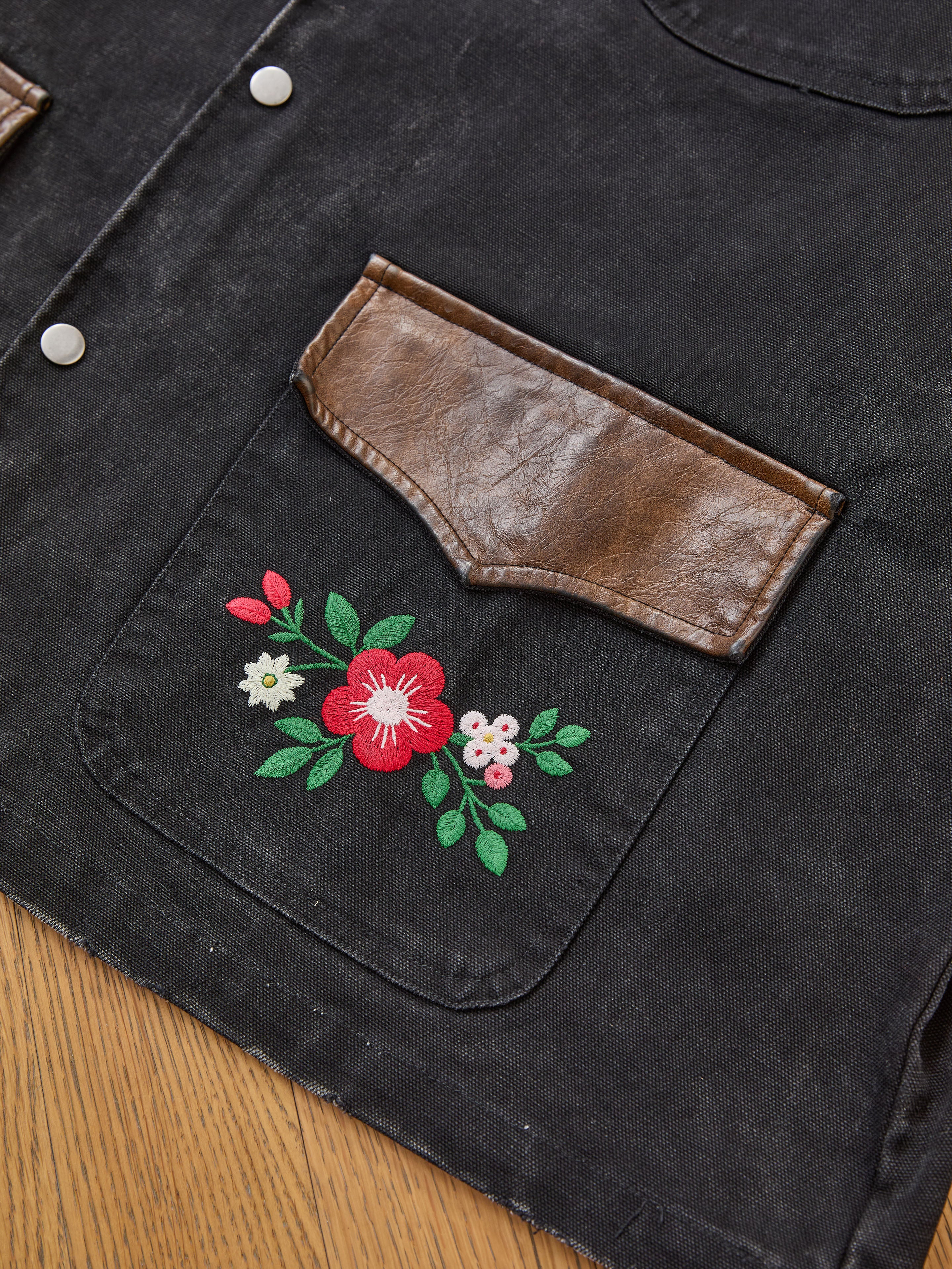 A close-up of the Found Canvas Embroidered Work Jacket features a black canvas design, brown leather pocket flap, and floral embroidery beneath the pocket, displayed on a wooden surface.
