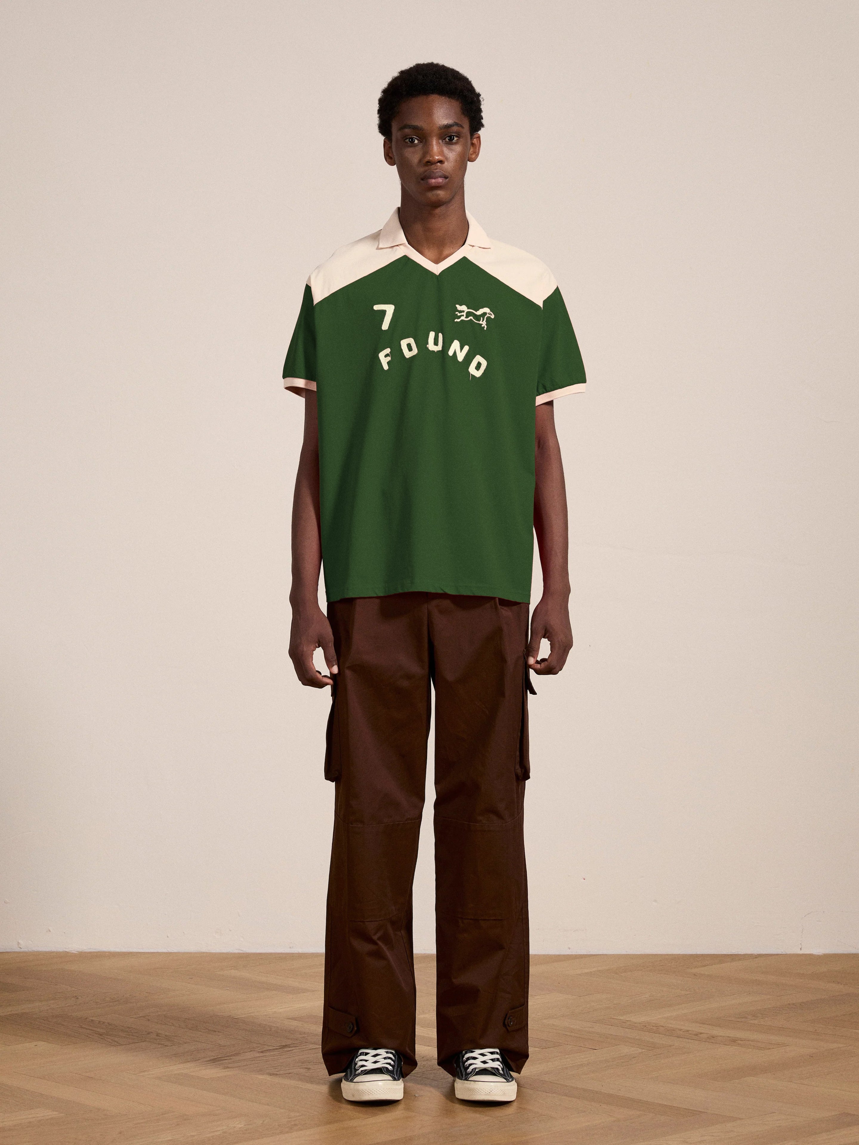 A person stands indoors on a wooden floor wearing the FOUND Goji Green Jersey, a green and white collared shirt with "7 FOUND" printed on it, paired with brown pants and black sneakers.