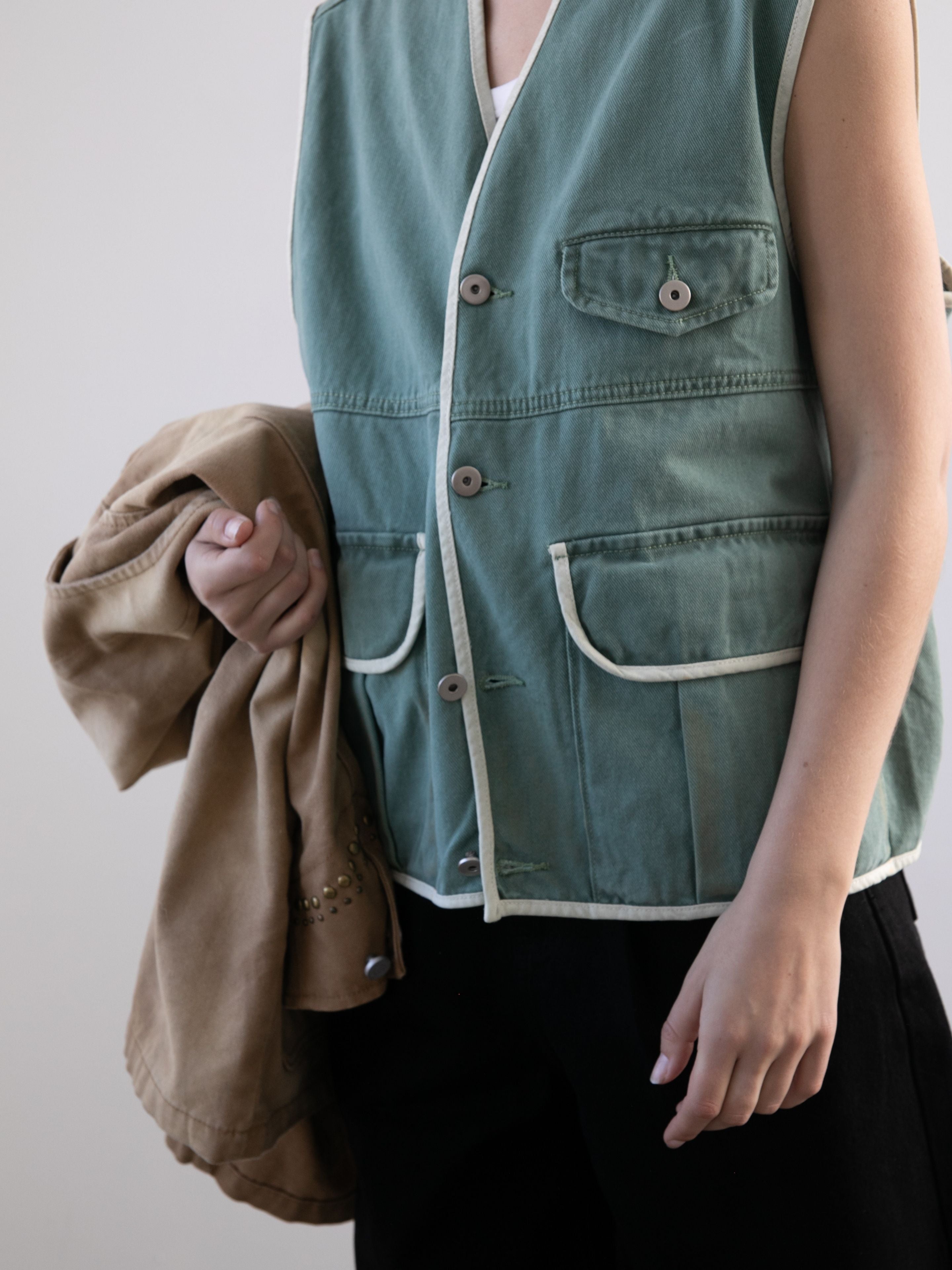 A person wears the FOUND Cotton Twill Utility Vest in vintage green over a white top and black pants, holding a tan jacket against a plain background.
