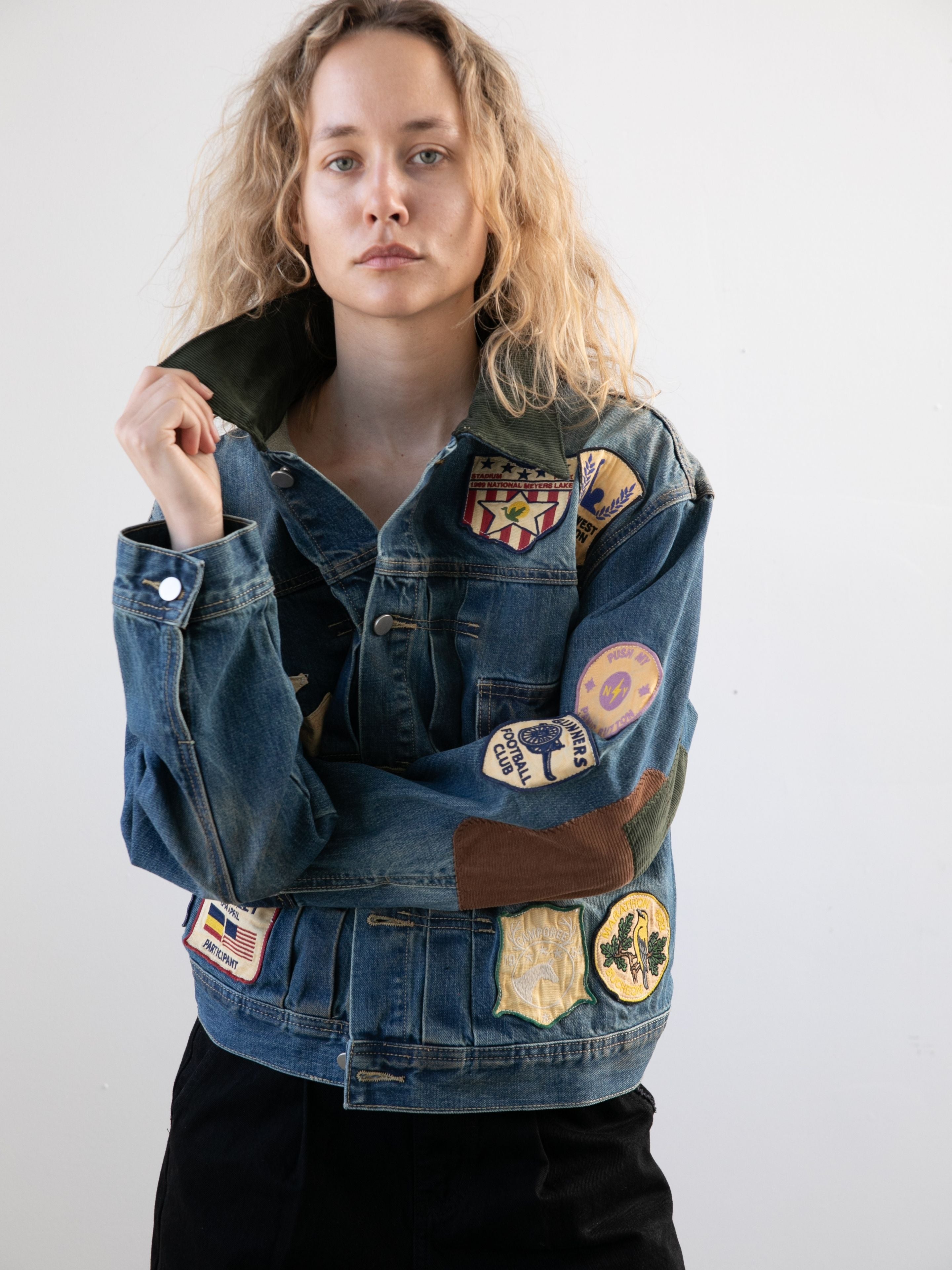 A person with wavy blonde hair models the FOUND Denim Patchwork Jacket, a unisex relaxed-fit piece featuring colorful patches and vintage washed denim, against a plain light background.