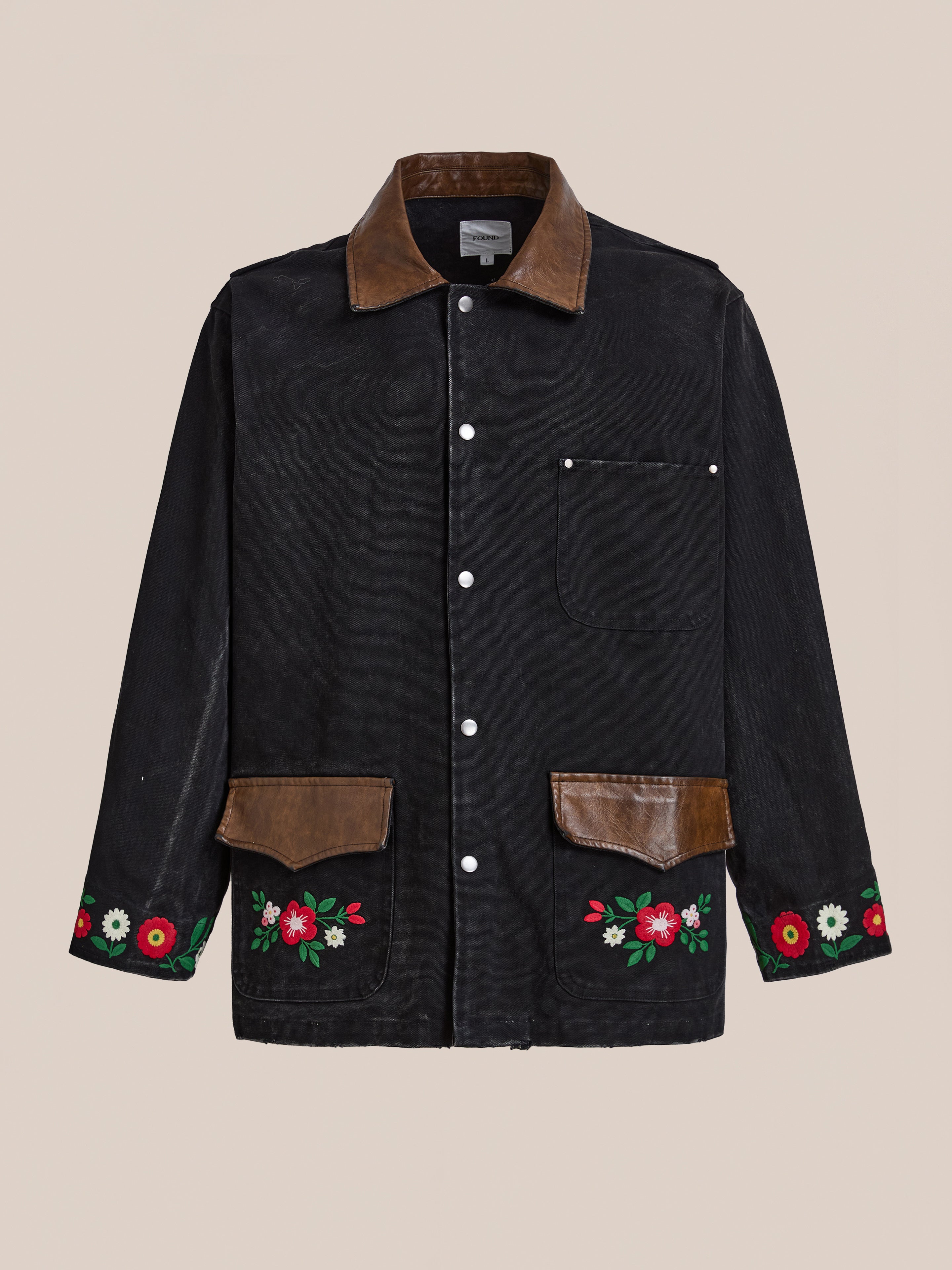 The Found Canvas Embroidered Work Jacket features a unisex relaxed fit in black canvas, brown leather collar and pocket flaps, front snap buttons, single chest pocket, and floral embroidery on the lower front and sleeves.