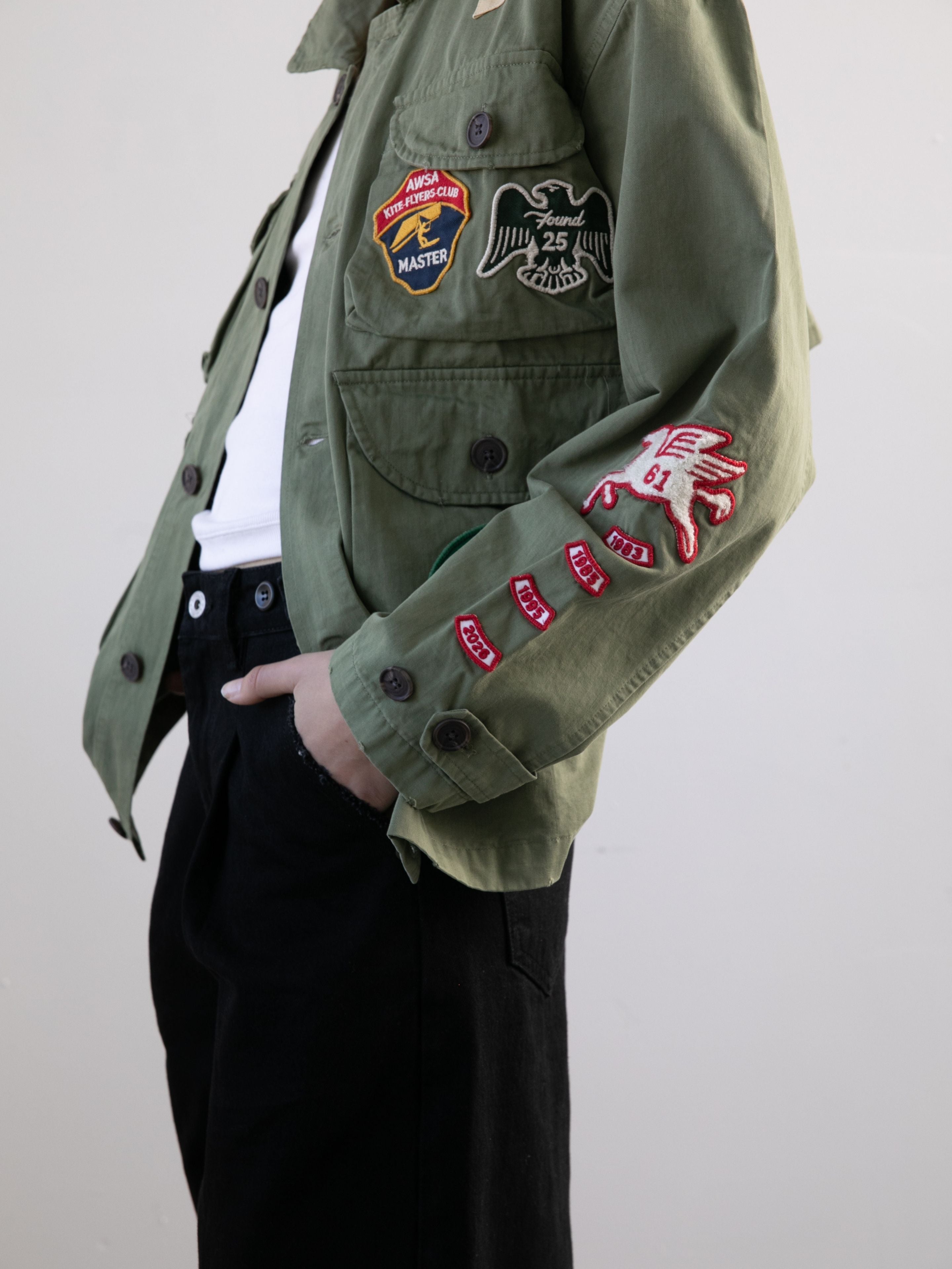 A person wears the FOUND Herringbone Utility Patchwork Jacket with embroidered patches and black pants, standing with one hand in their pocket.