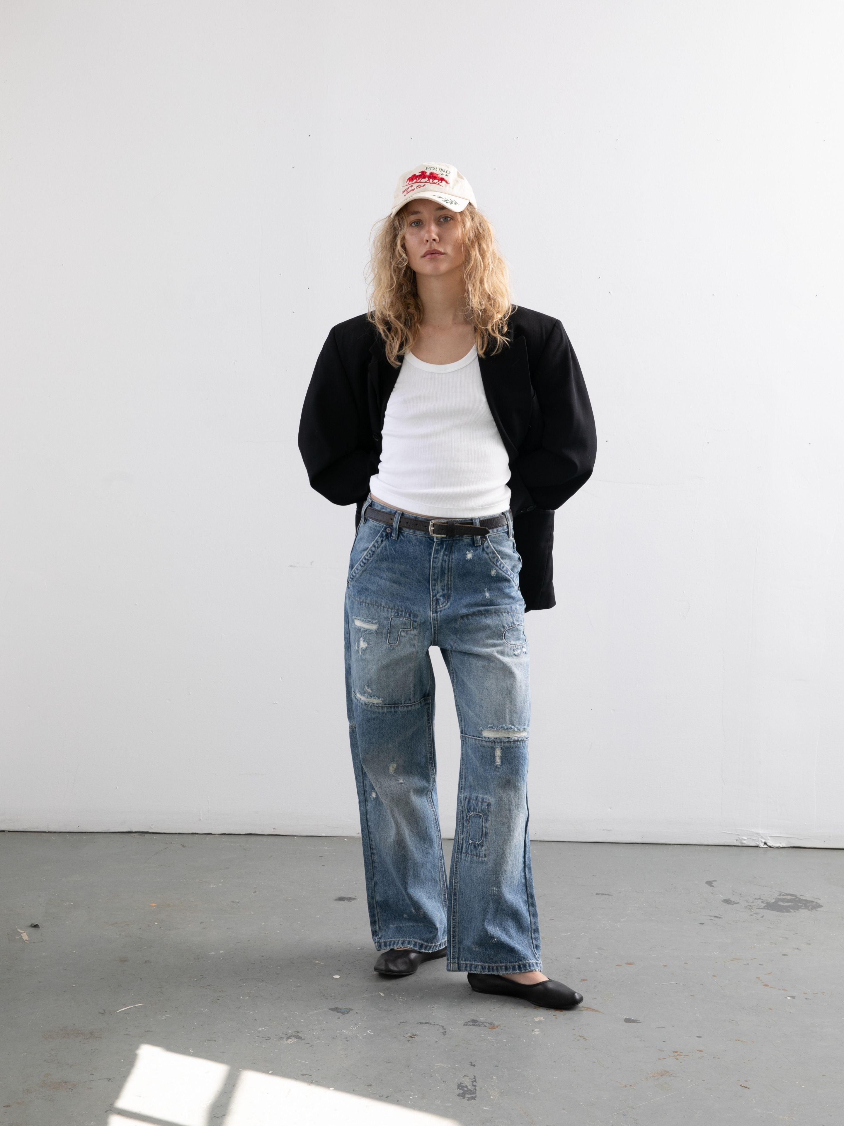 A person wearing a white cap, black jacket, white tank top, and FOUND Painter Repaired Jeans stands against a plain white wall.