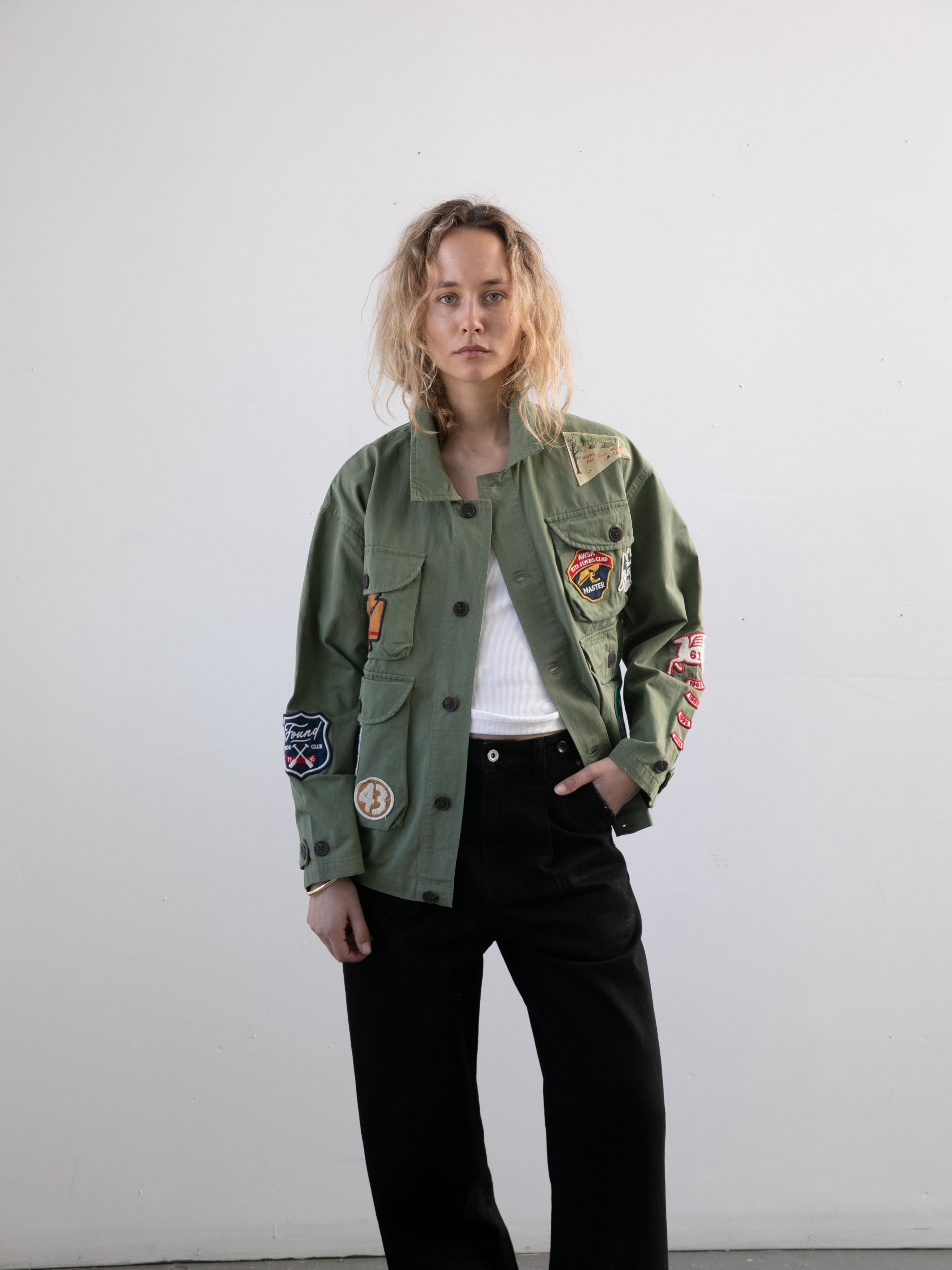 A person with wavy blonde hair wears the FOUND Herringbone Utility Patchwork Jacket over a white shirt and black pants, standing against a plain light-colored wall.