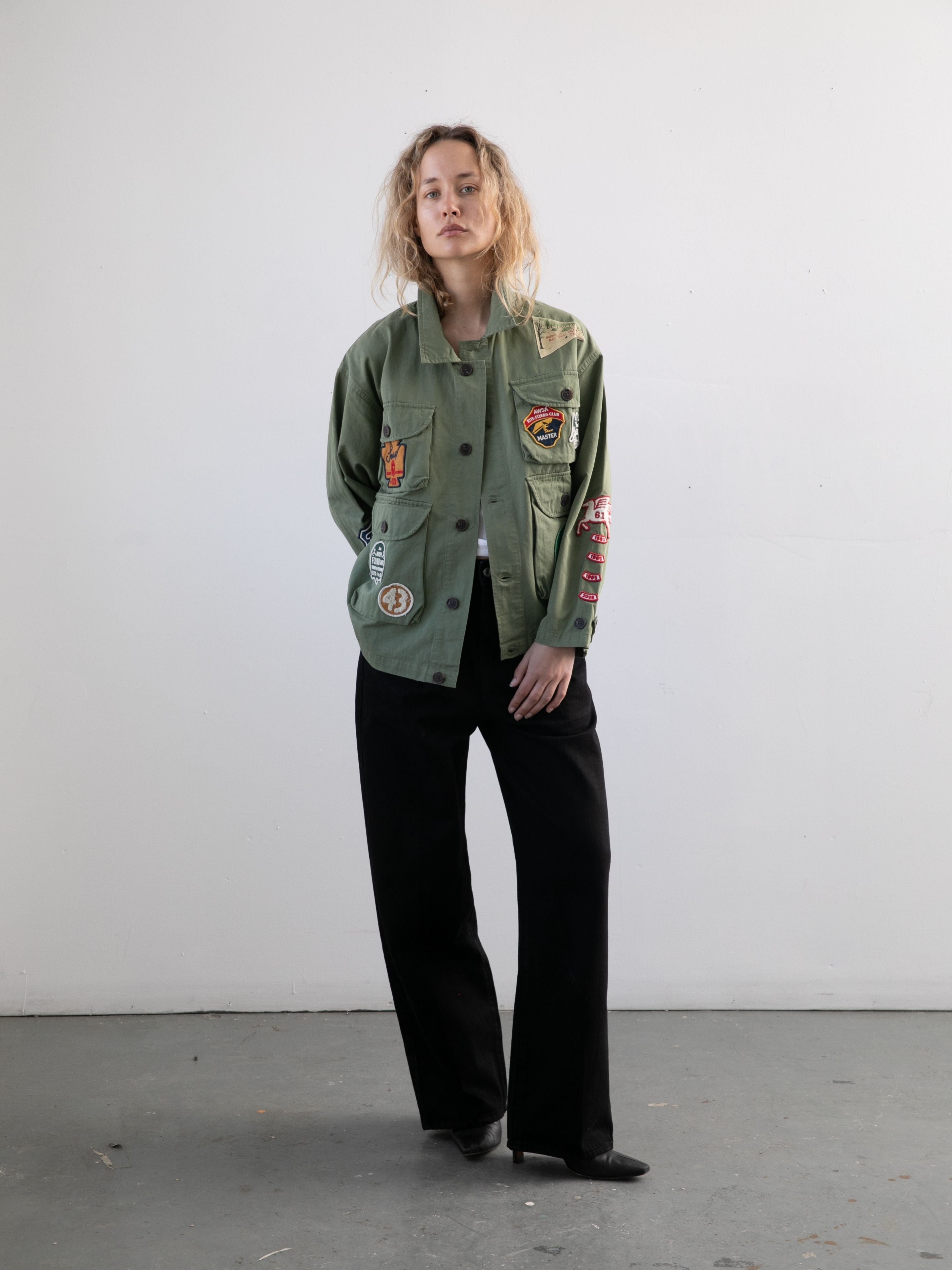 A person stands indoors against a plain white wall, wearing the FOUND Herringbone Utility Patchwork Jacket over a black outfit with black shoes.