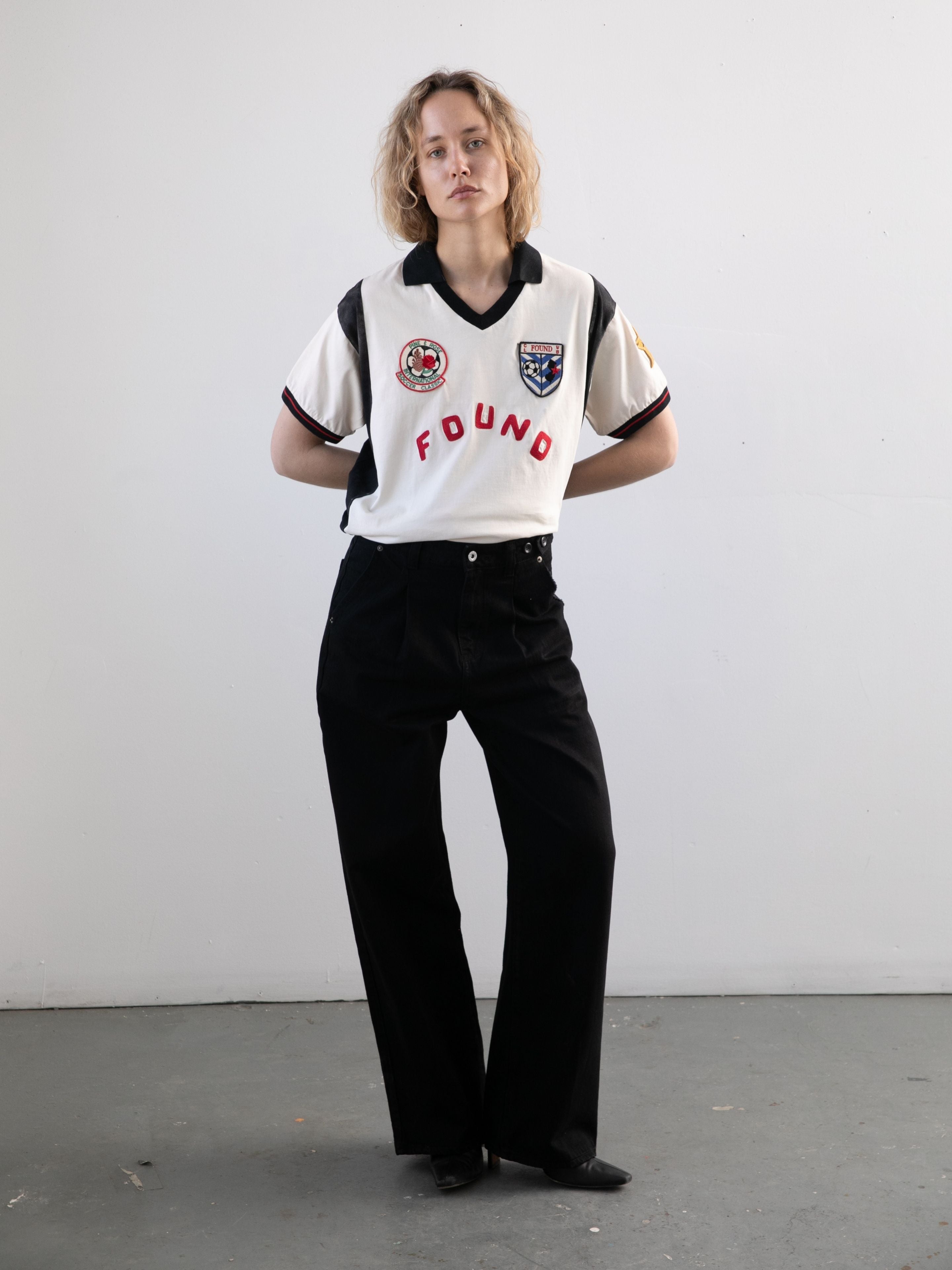 A person stands indoors by a plain wall, wearing the FOUND Union Soccer Jersey—styled like a vintage football shirt—with black pants and black shoes.