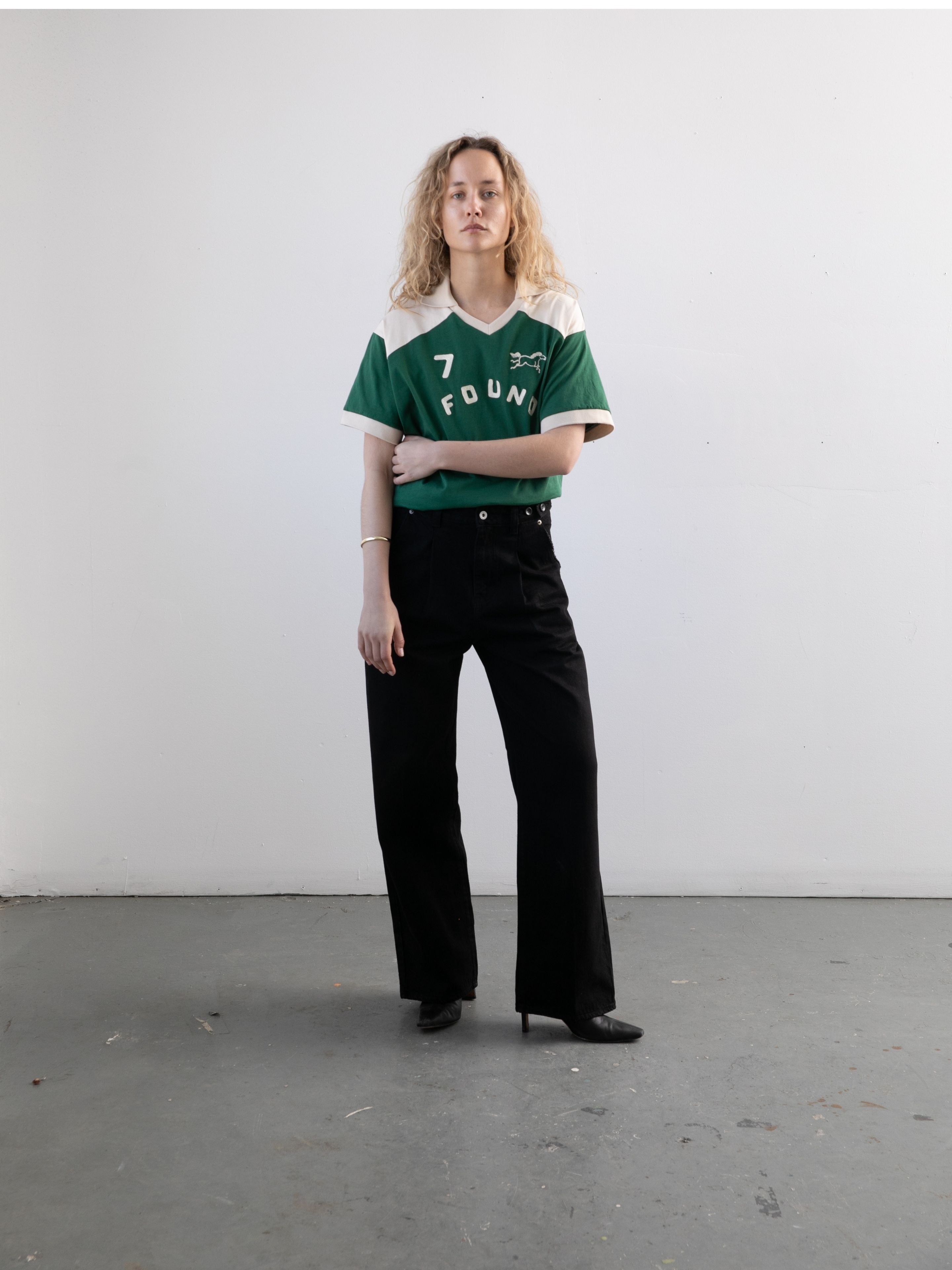 A person with curly hair stands indoors against a plain white wall, wearing the FOUND Goji Green Jersey, paired with black pants and black shoes.