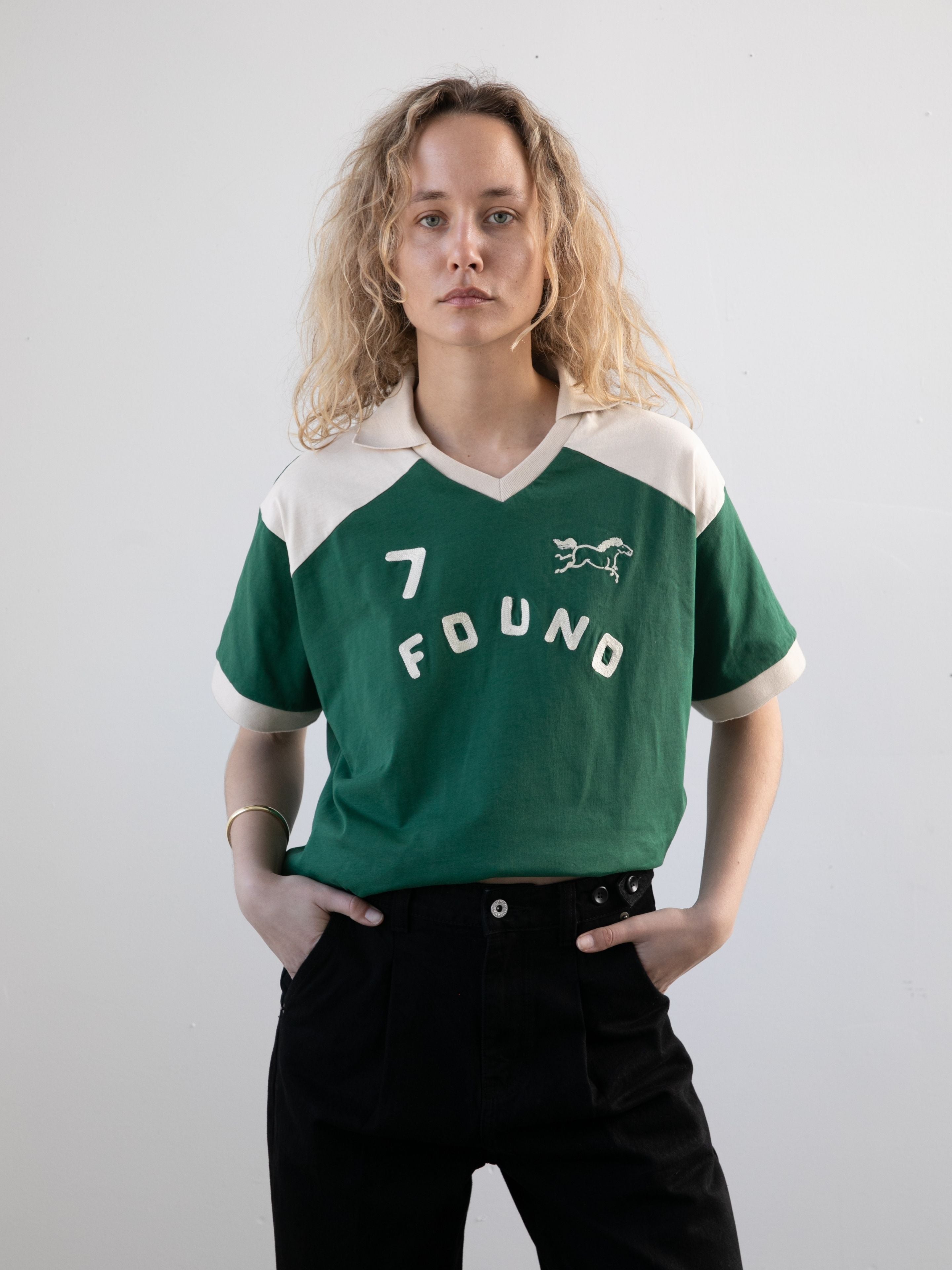 A person with wavy blonde hair wears the FOUND Goji Green Jersey and black pants, standing against a plain light background.