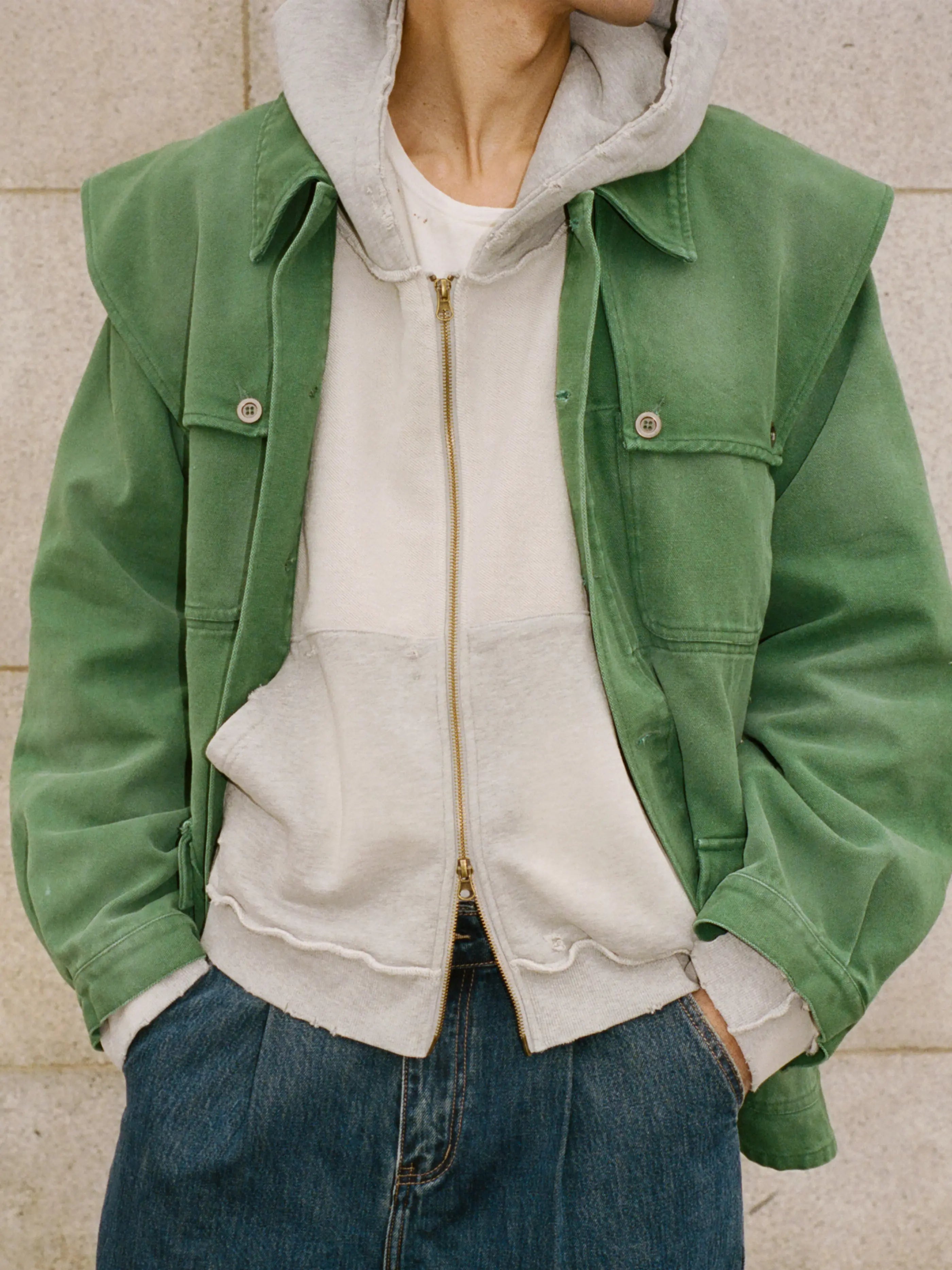 A person wears the FOUND Deconstructed Terry Zip Up Hoodie over a white T-shirt, paired with blue jeans, and stands against a light stone wall.