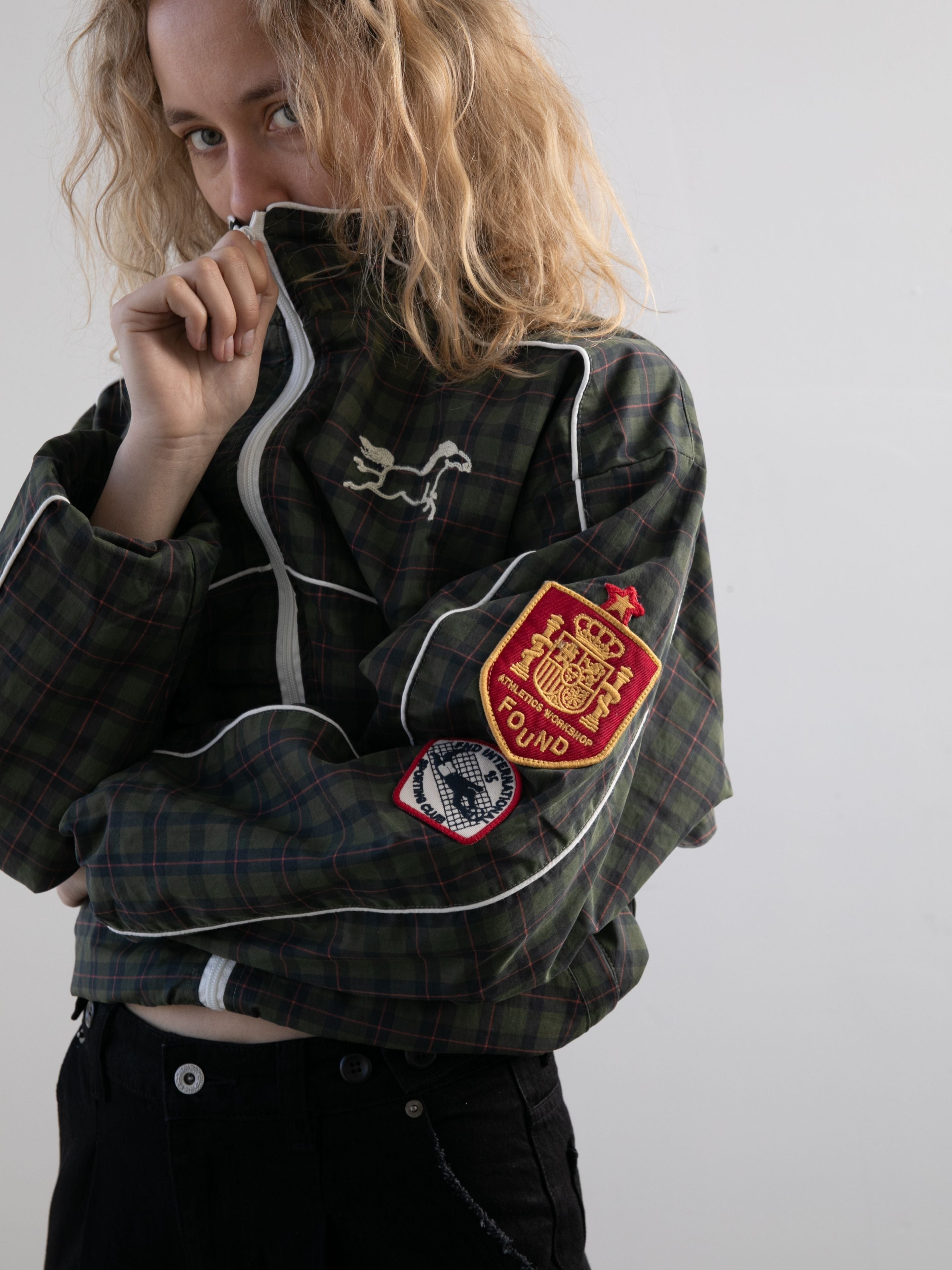 A person with curly blond hair partially covers their face with the FOUND Forest Plaid Track Jacket, which features several embroidered patches on the sleeve.