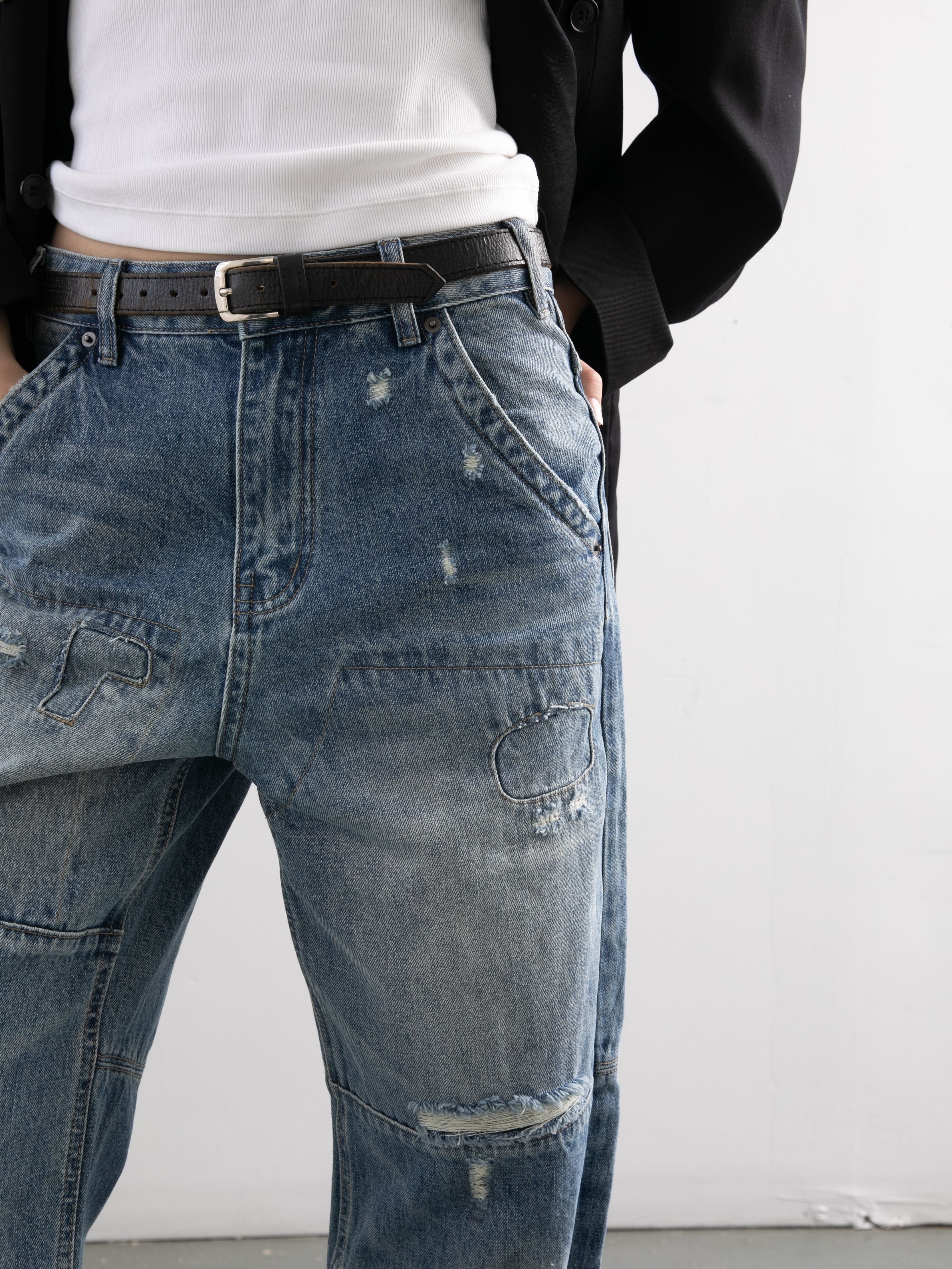 A person wearing FOUND Painter Repaired Jeans, a black belt, white shirt, and black jacket stands with hands in pockets against a plain light background.