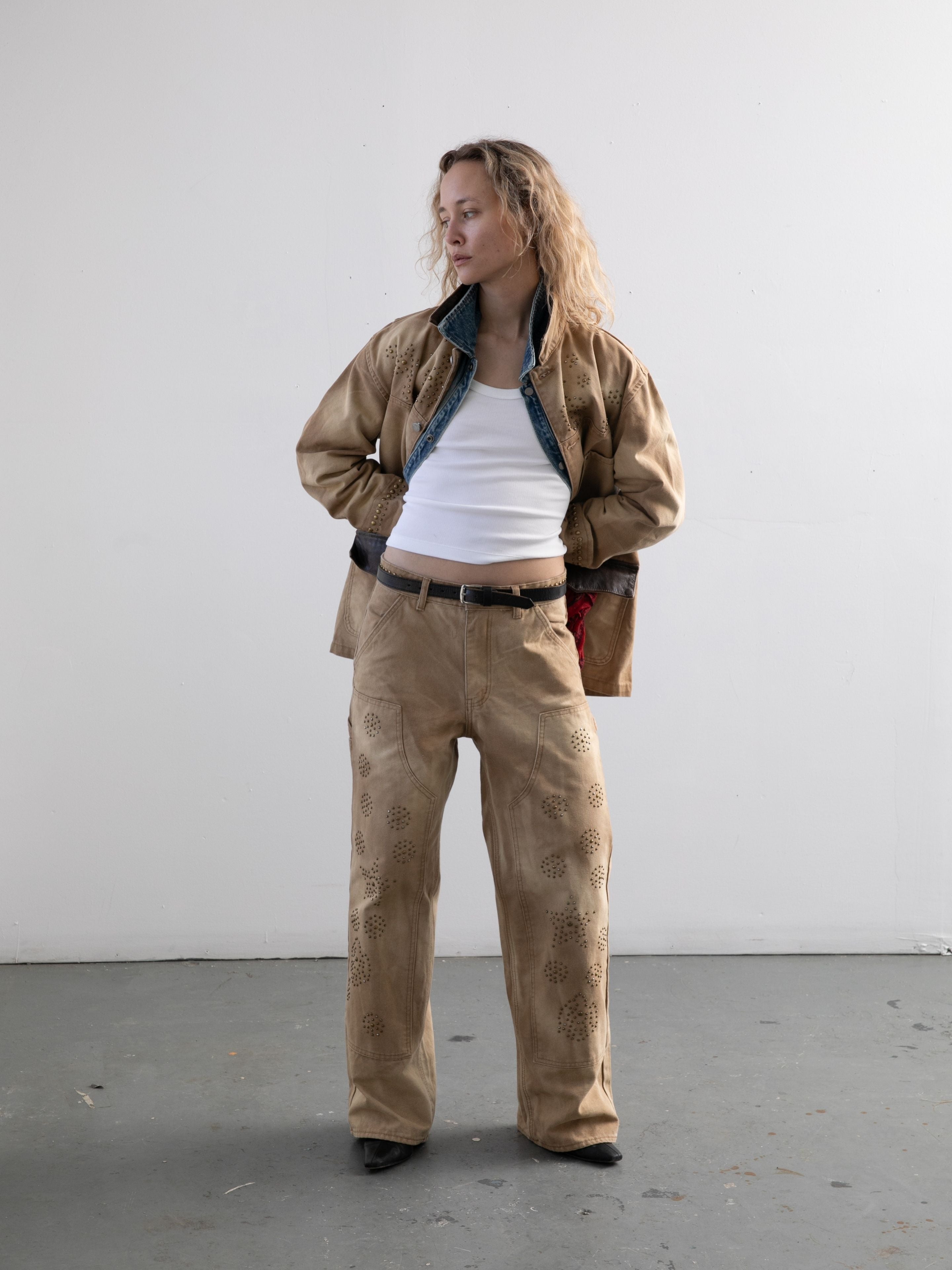 Indoors, a person wears the Found Canvas Studded Barn Jacket and matching pants over a white tank top, hands in the jacket pockets and looking to the side.