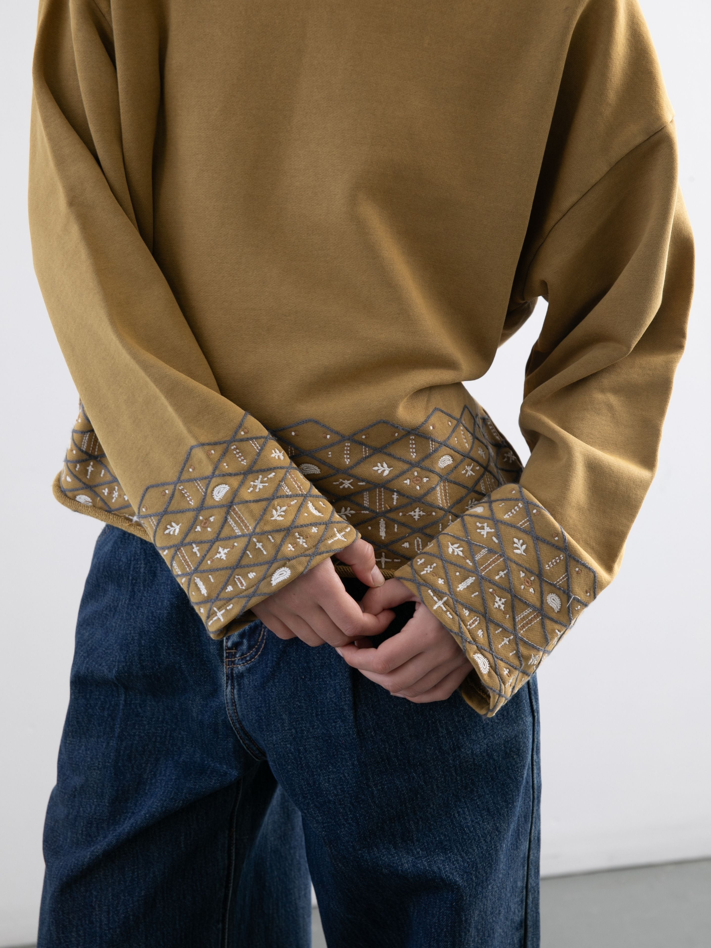 Someone wears the FOUND Embroidered Border Hoodie in washed dark olive, featuring patterned cuffs, paired with blue jeans and stands with hands clasped in front.