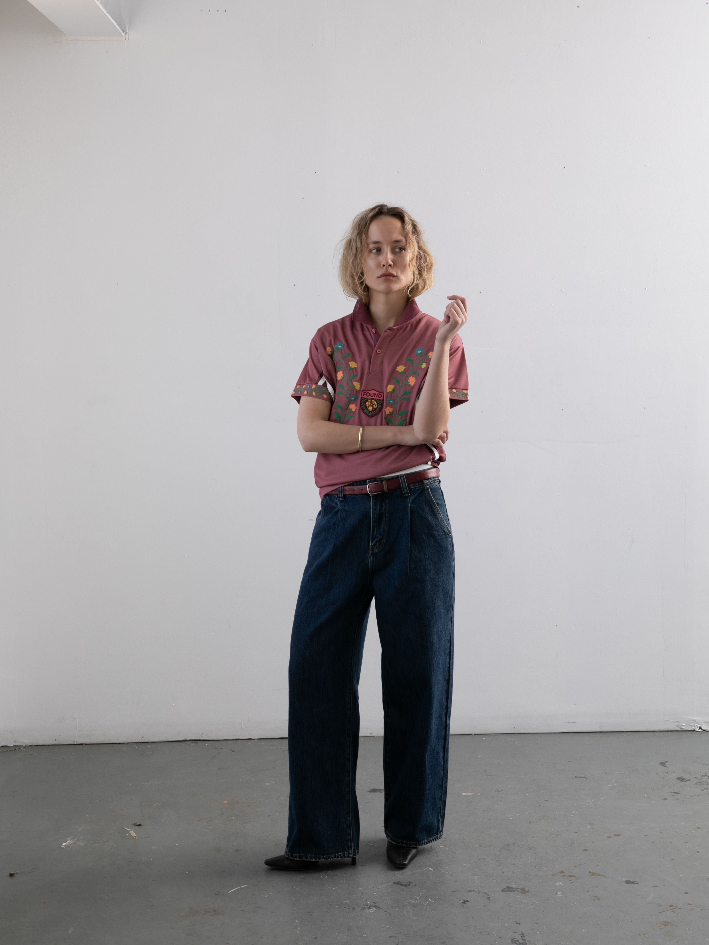 A person with short blonde hair models the FOUND Floral Polo Jersey, paired with wide-leg jeans, standing against a plain white wall and looking at the camera with one arm crossed.