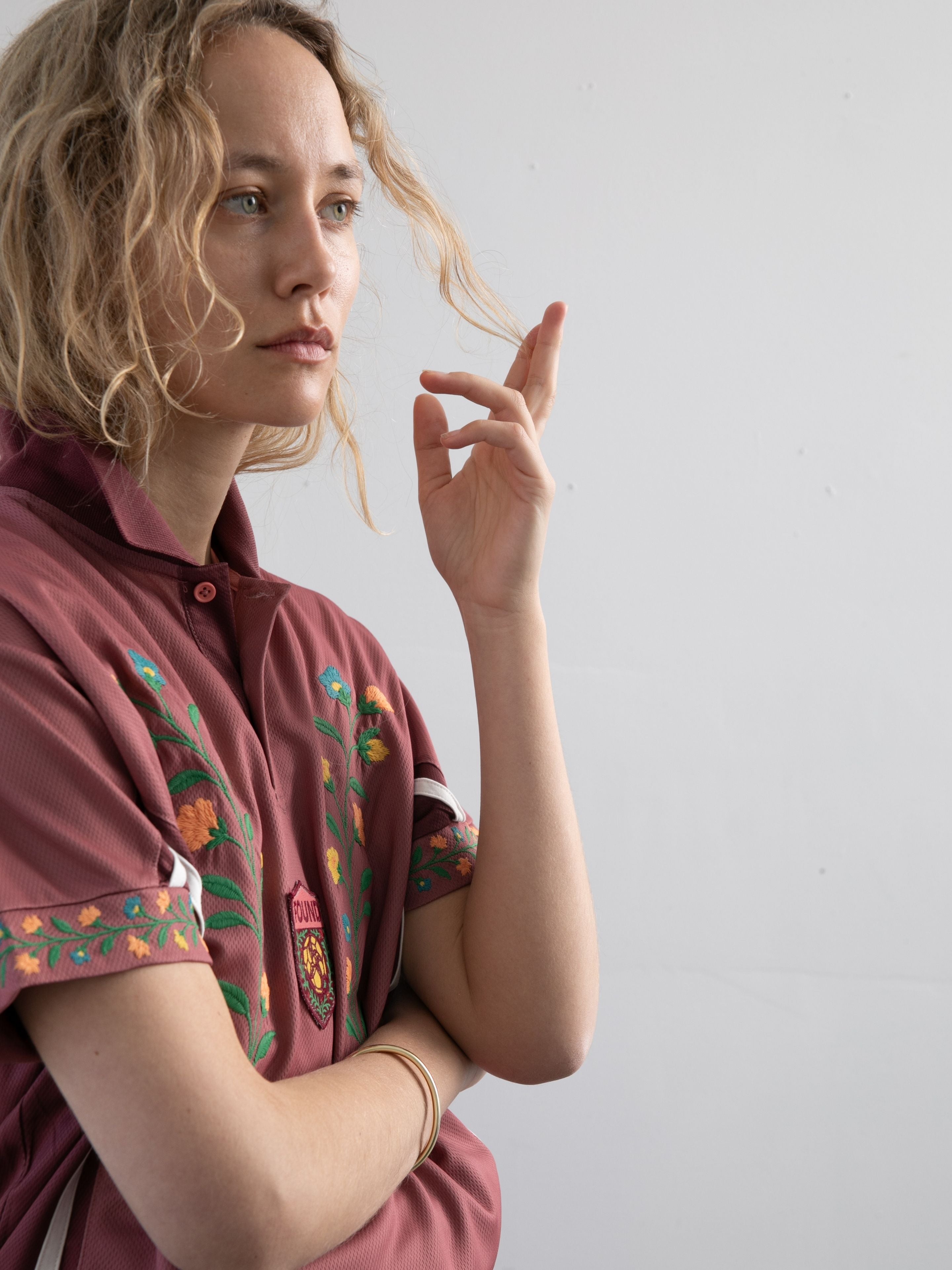 A person with wavy blonde hair wears the FOUND Floral Polo Jersey, standing against a plain light wall and gazing thoughtfully to the side.