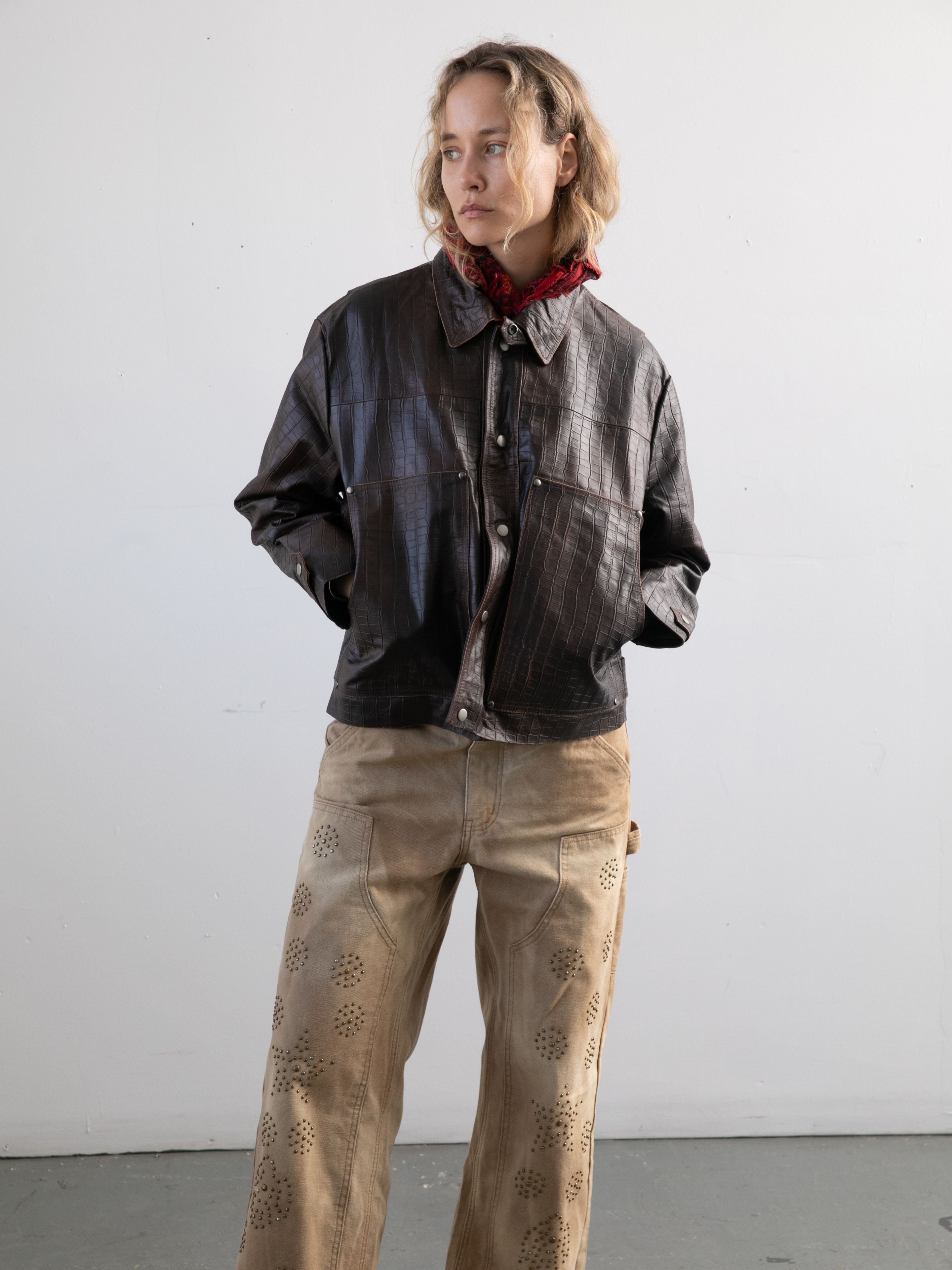A person stands against a plain wall, wearing the FOUND Distressed Croc Embossed Leather Work Jacket, beige patterned pants, and a red scarf, with hands tucked into the jacket pockets.