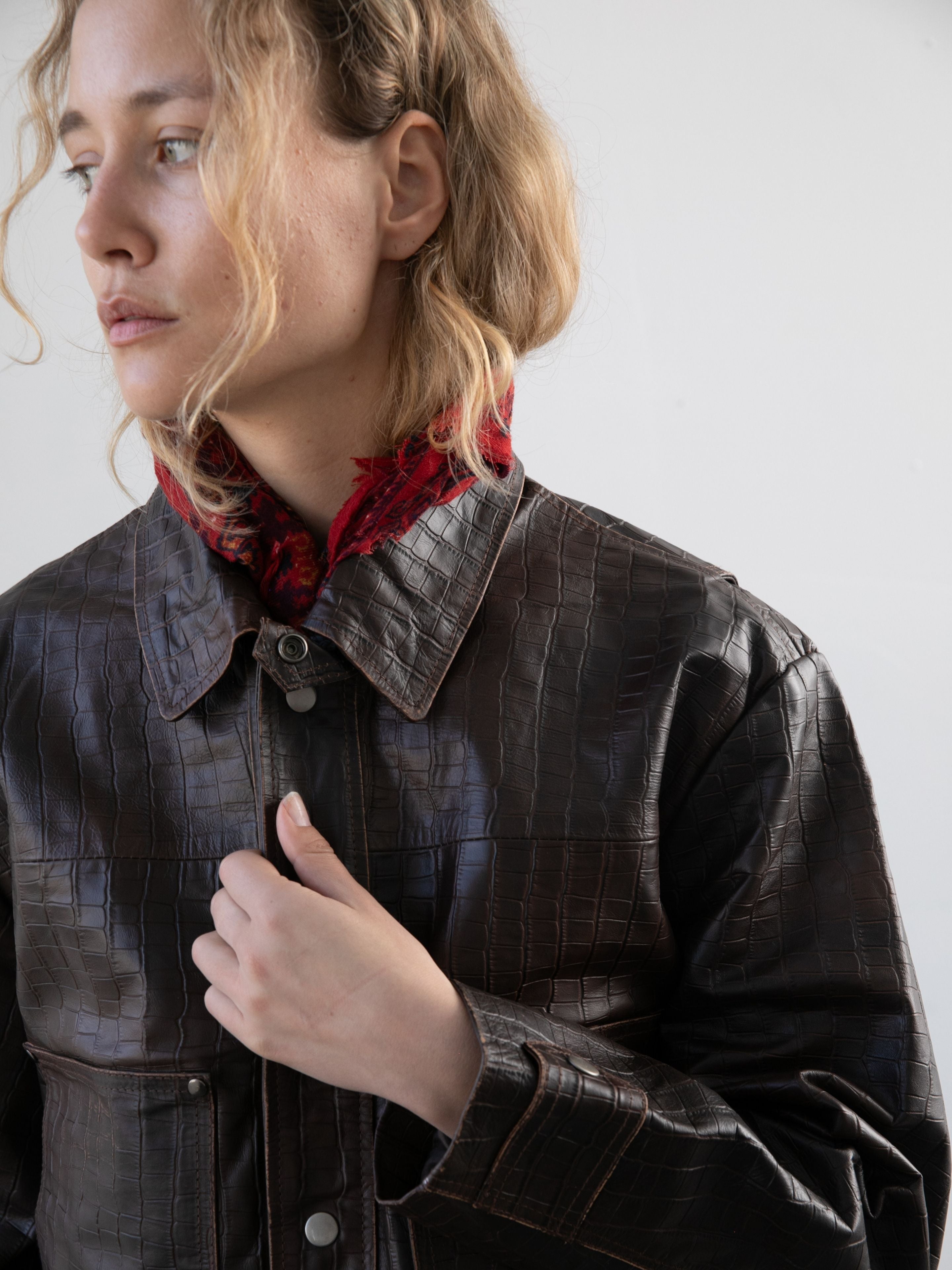 A person with wavy blonde hair wears the FOUND Distressed Croc Embossed Leather Work Jacket and a red patterned scarf, gazing to the side against a plain background.