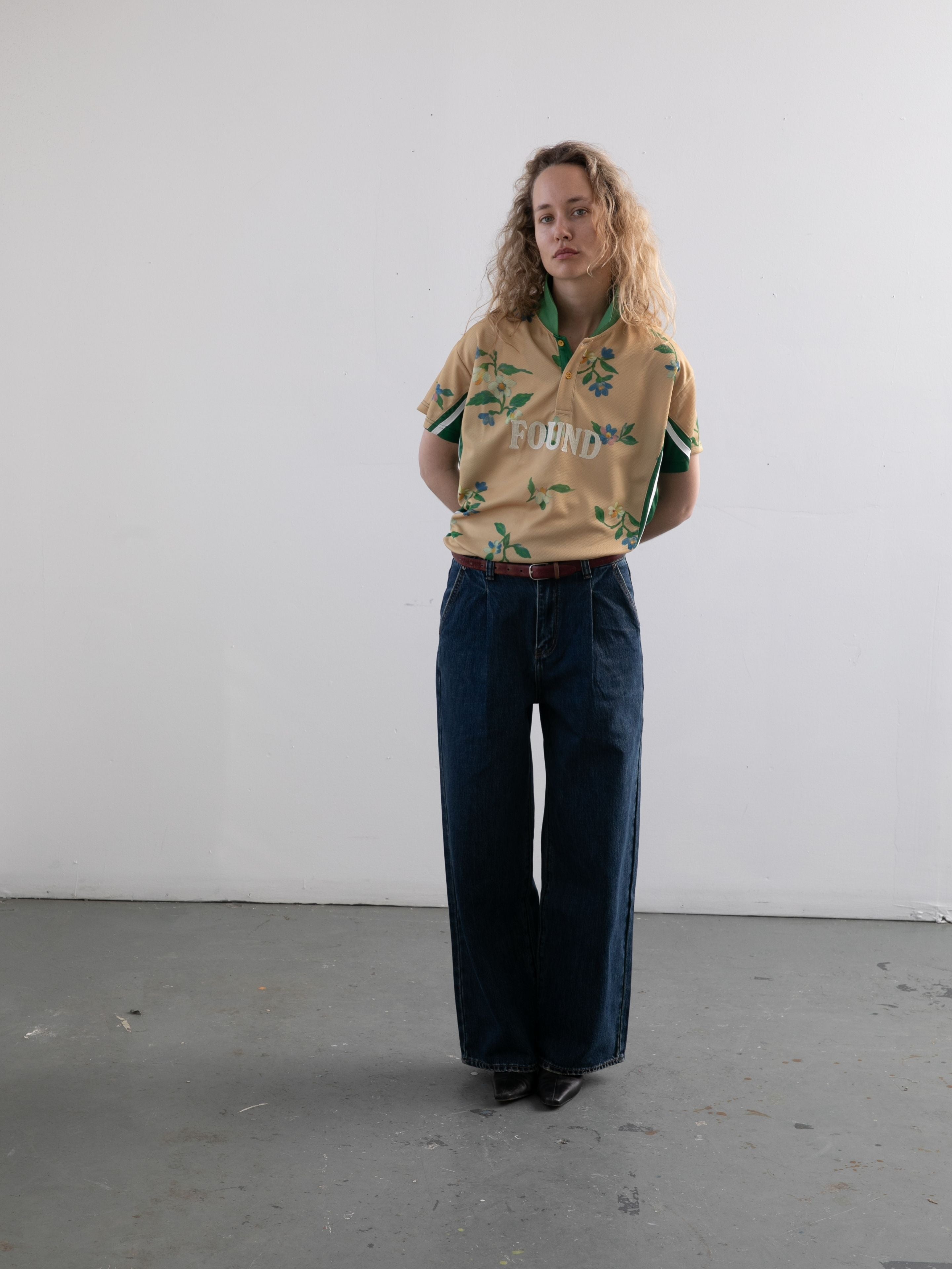 A person with curly hair stands against a plain white wall, wearing FOUND's Floral Polo Jersey and dark wide-leg jeans.