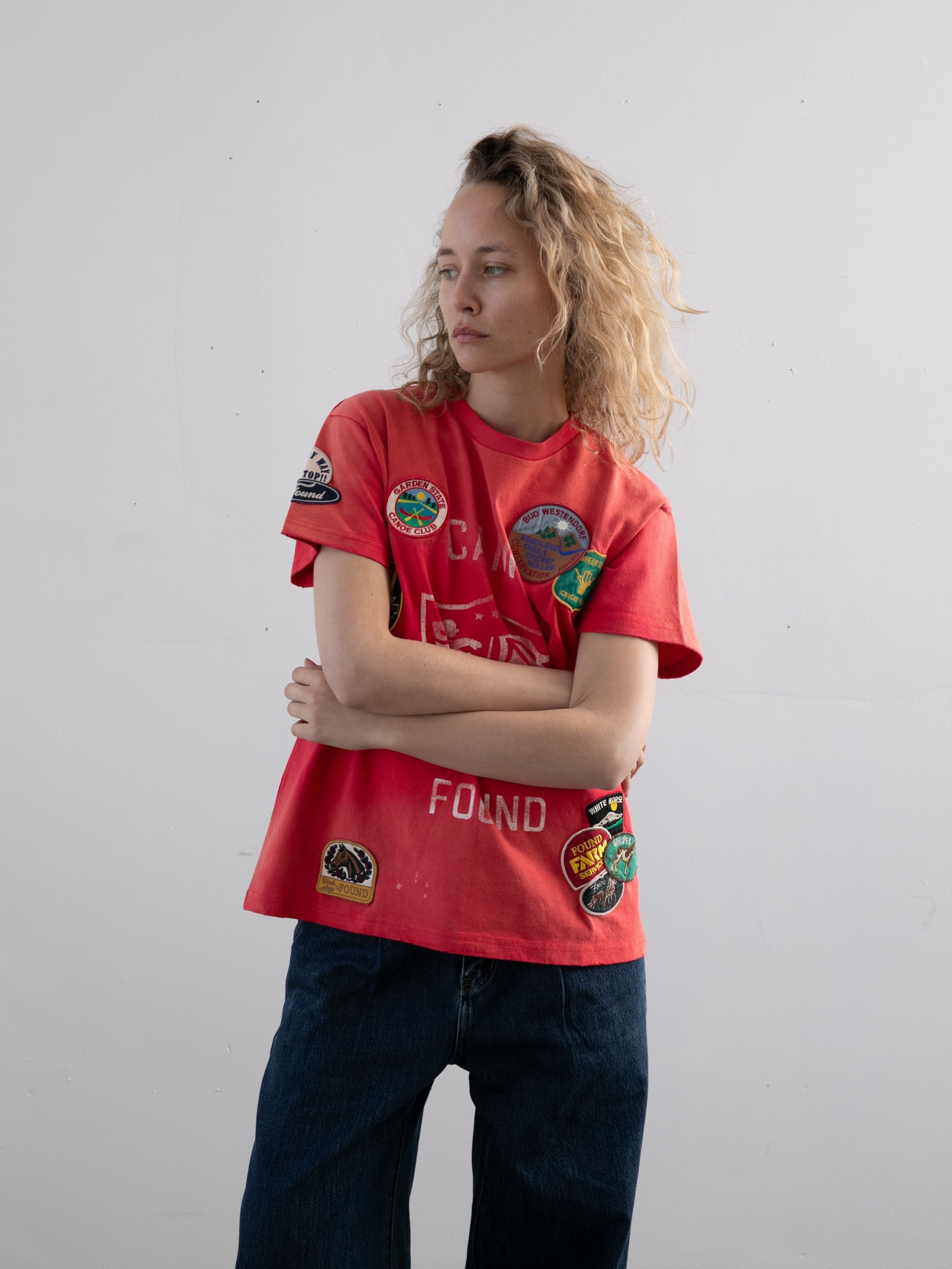 A person with curly blonde hair wears the FOUND Patchwork Camp Tee in red and dark blue jeans, standing with arms crossed against a plain white background.