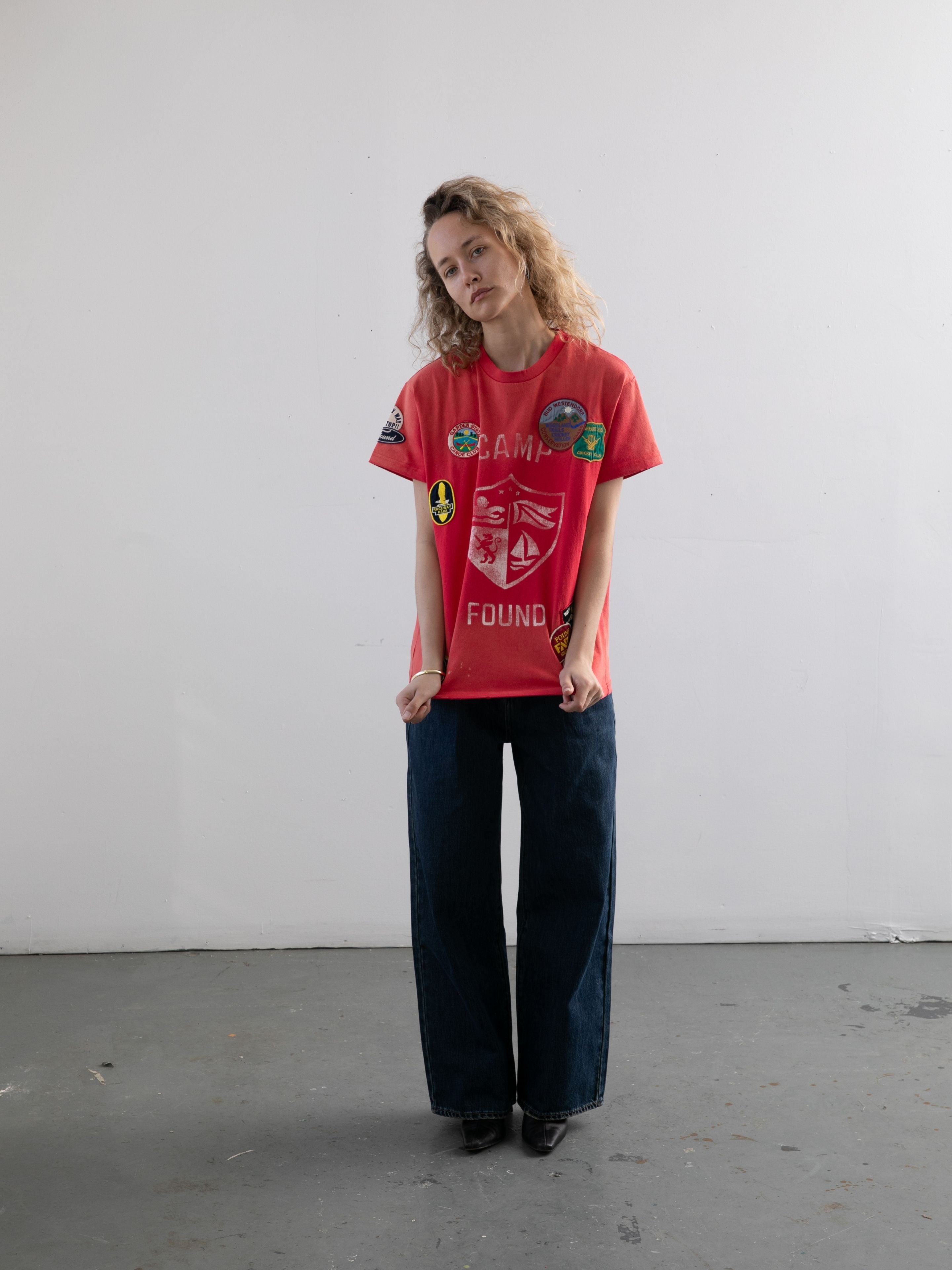 A woman with curly hair wears the FOUND Patchwork Camp Tee in red and dark wide-leg jeans, standing against a plain white wall on a gray floor.