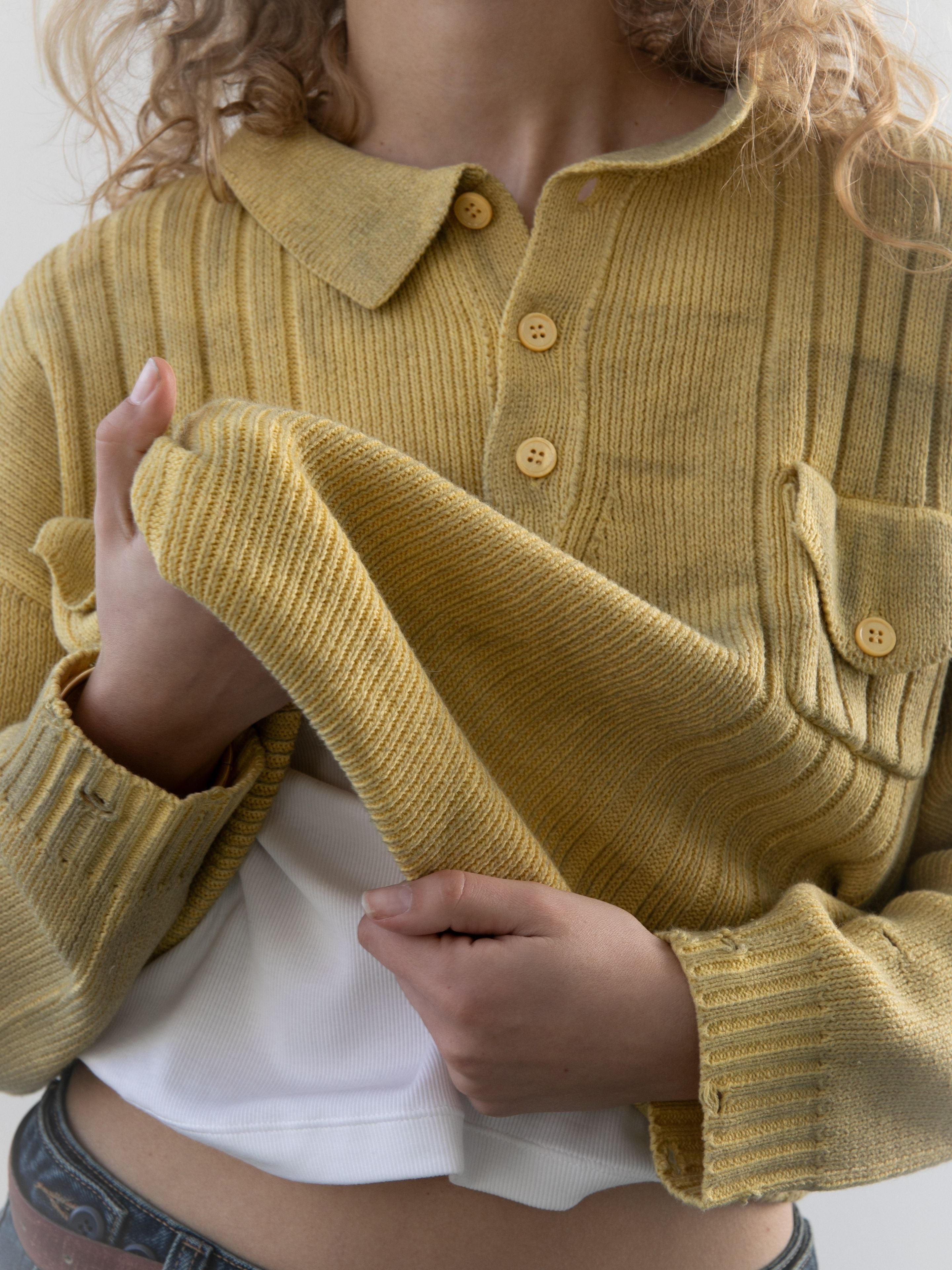 A model lifts part of the FOUND Distressed Pullover Knit, showing a white shirt beneath. With only the torso and one hand visible, this yellow ribbed unisex sweater creates an easy transitional weather style.