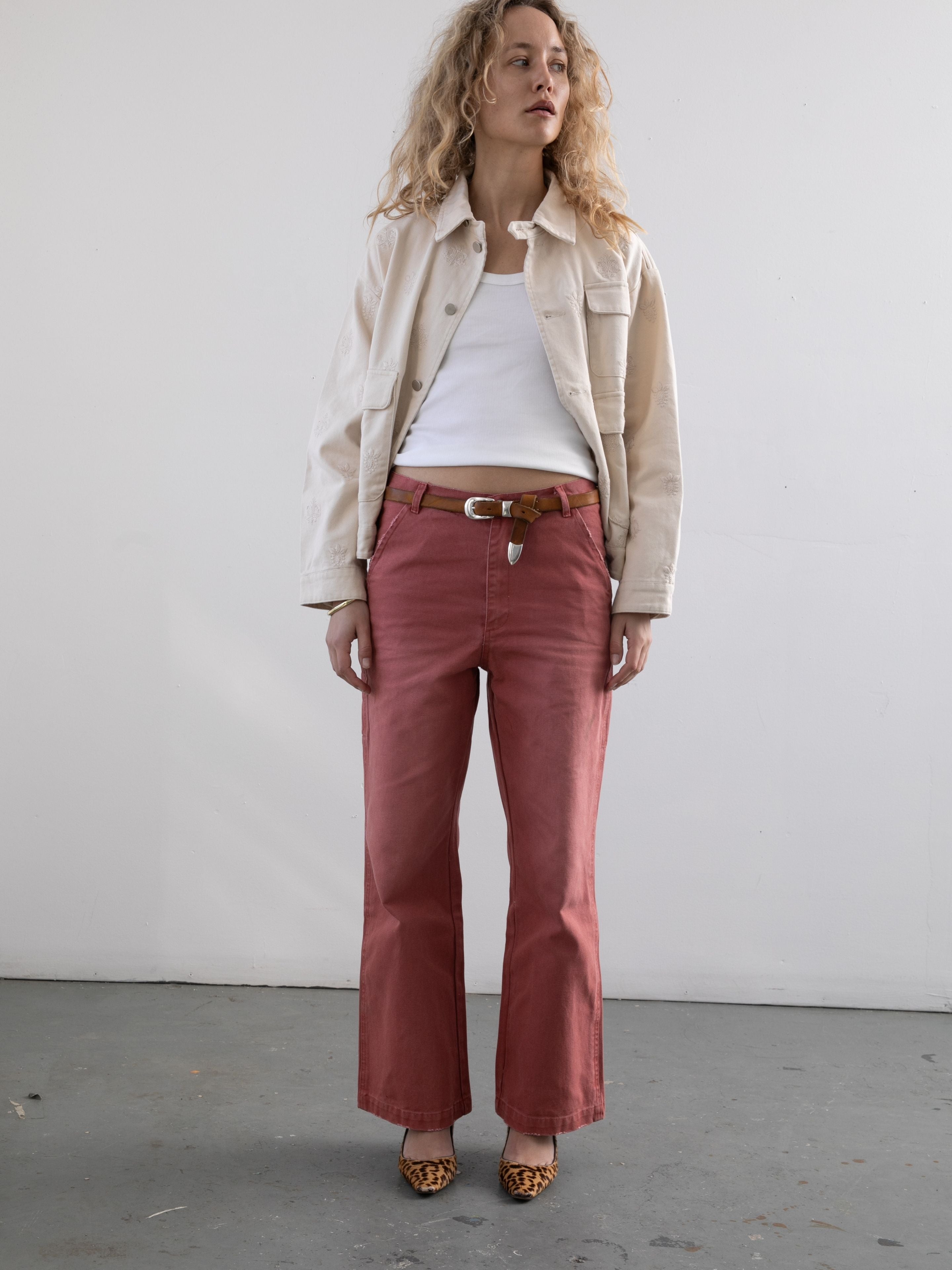 A person stands against a plain wall wearing Found's Embroidered Motif Jacket, a white tank top, red pants with a brown belt, and leopard print heels.