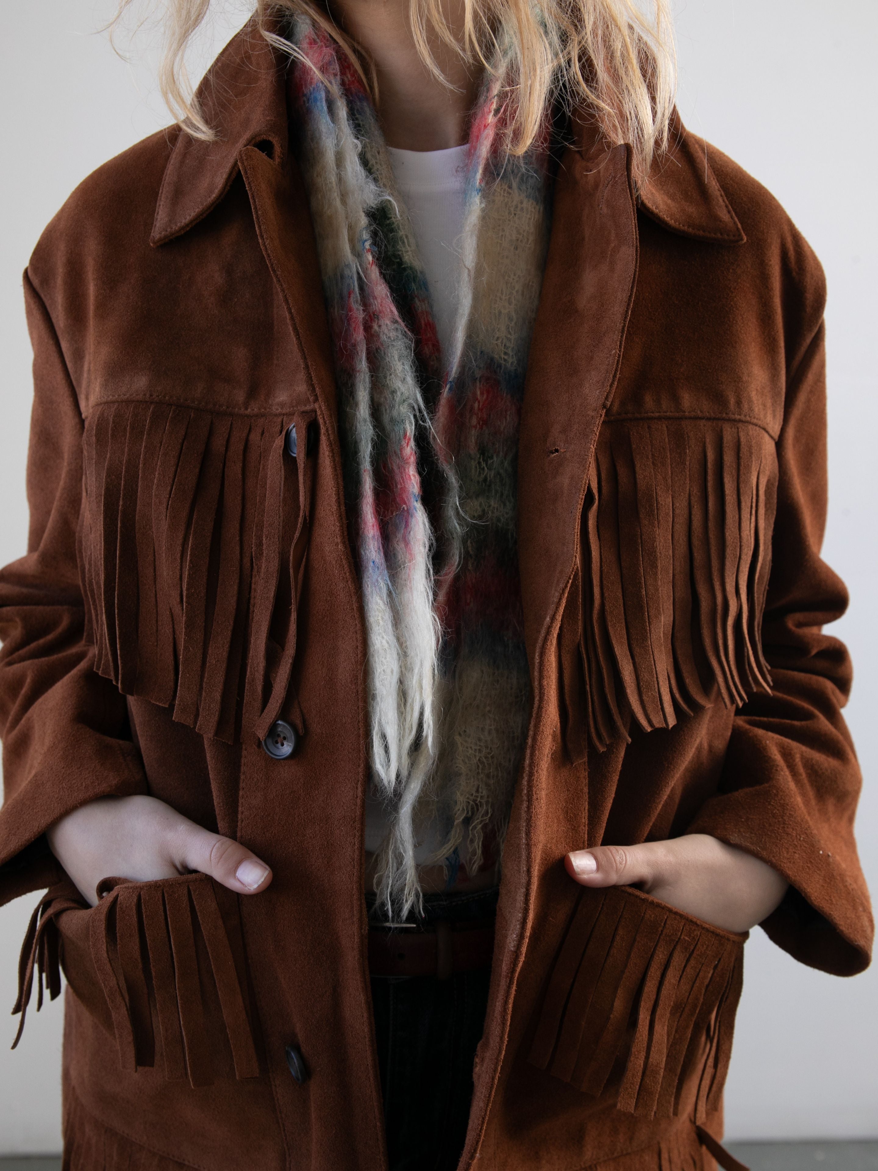 A person wears the FOUND Western Suede Fringe Jacket and a multicolored fuzzy scarf, hands in pockets, standing against a plain background.