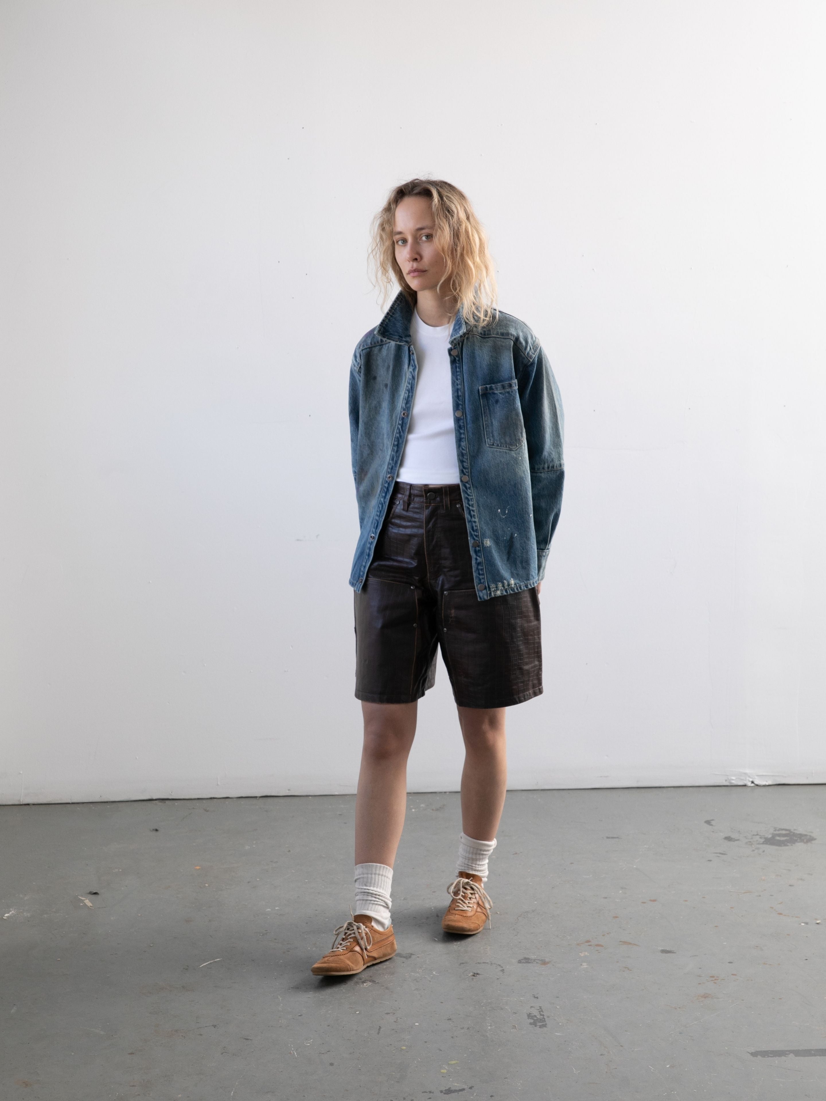 Indoors against a white wall, a person wears a blue denim jacket, white t-shirt, FOUND Distressed Croc Embossed Leather Shorts, tan shoes, and white socks.