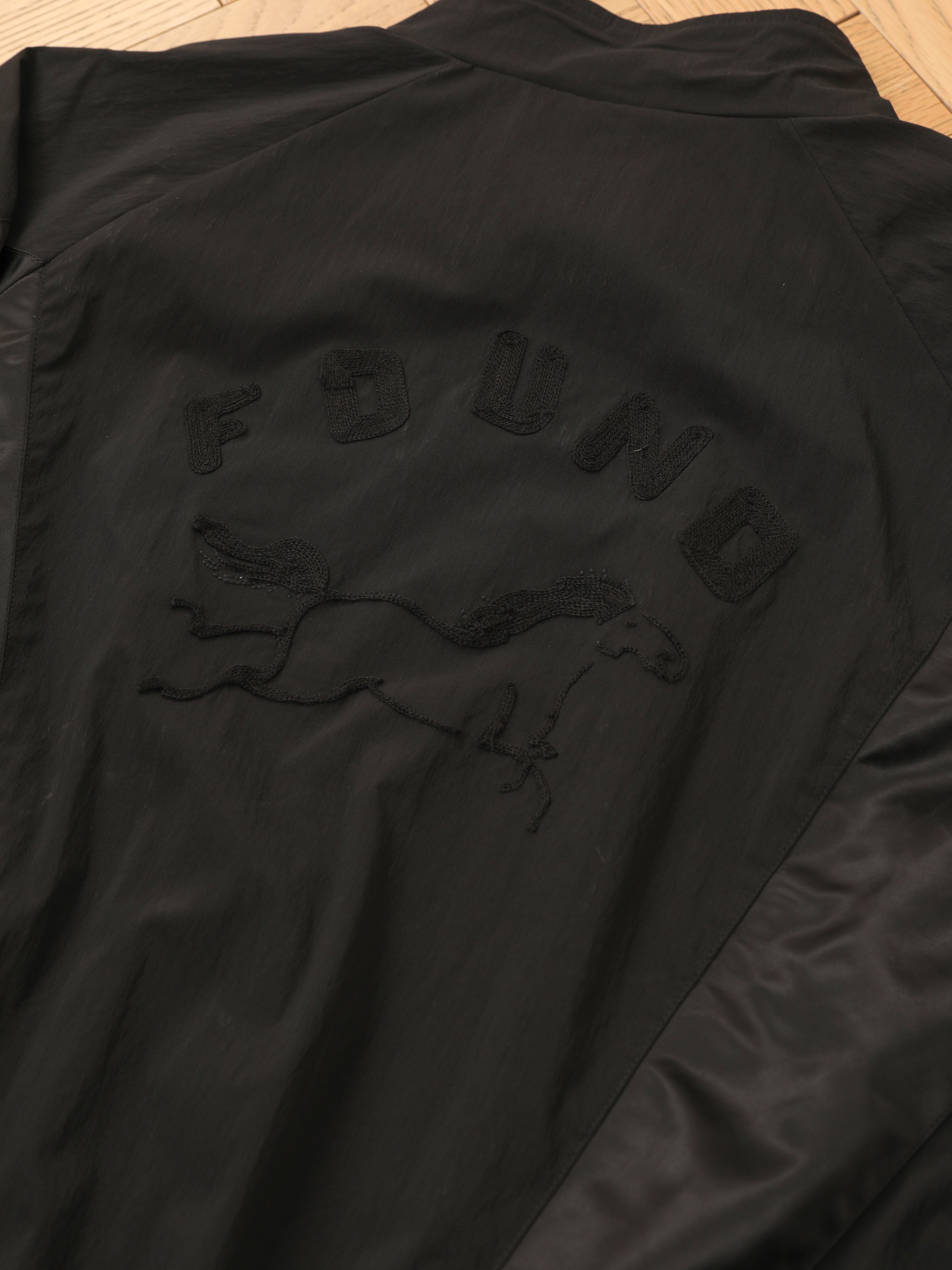 The FOUND Pine and Rose Jacket, a black vintage sportswear track jacket, is laid flat on wood with "FOUND" and a horse graphic embroidered on the back.