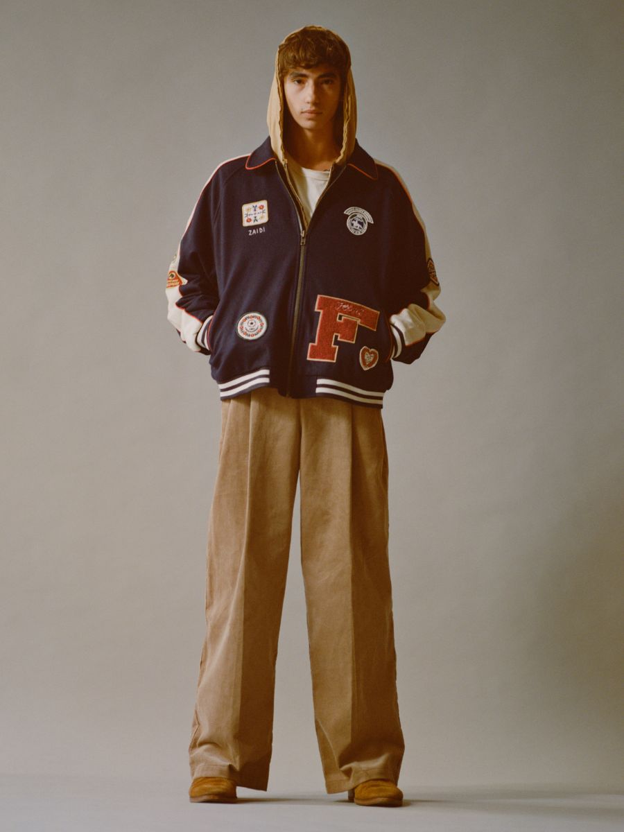 Sporting a vintage look, a person wears the All Roads Varsity Jacket from FOUND, complete with patches and a hood. They pair it with a light shirt, loose beige pants, and brown shoes against a plain backdrop.