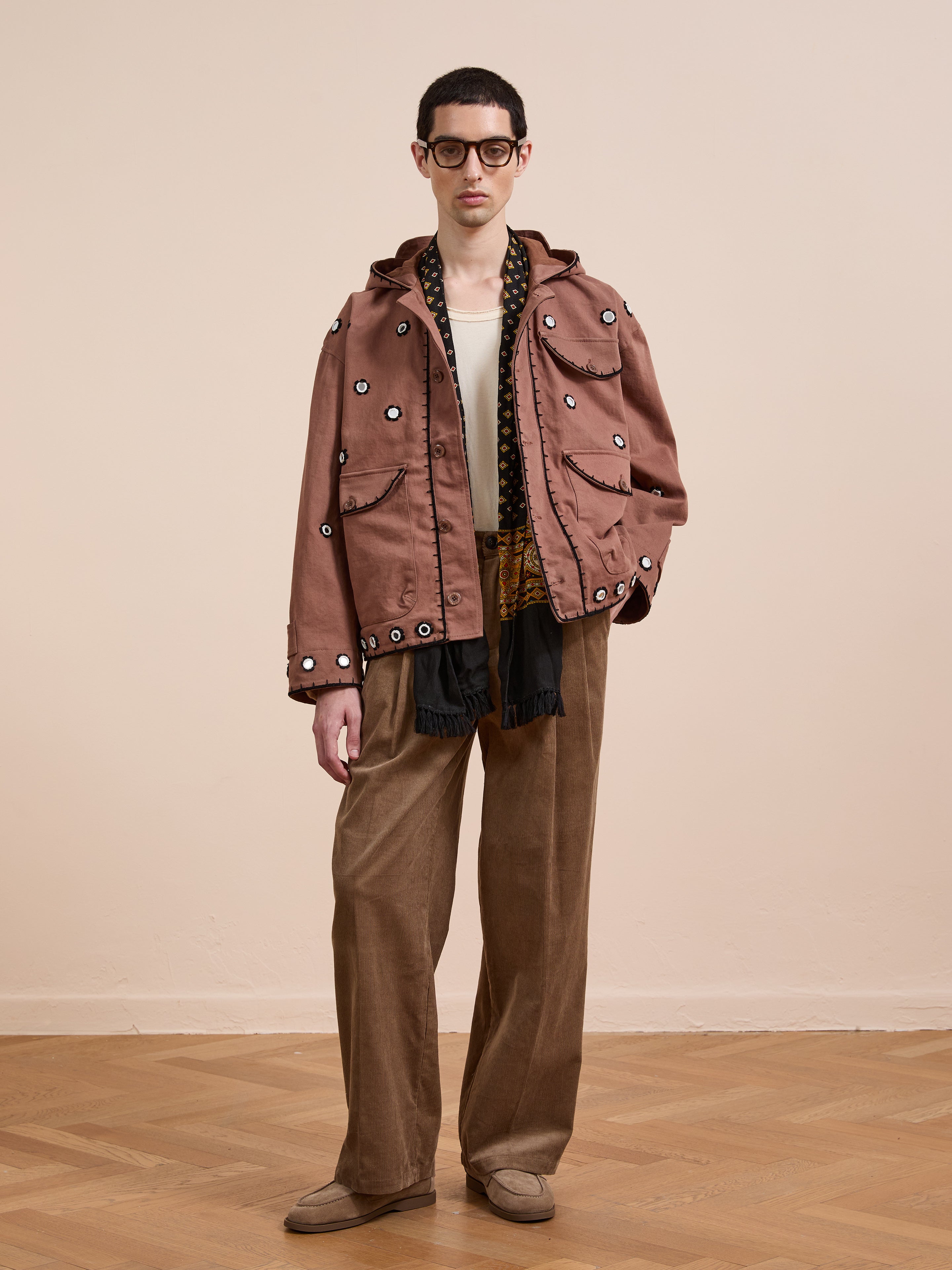 Indoors, a person wears FOUND's Hand Embroidered Hooded Mirror Anorak, wide-legged brown pants, a white shirt, patterned scarf, brown shoes, and dark sunglasses.