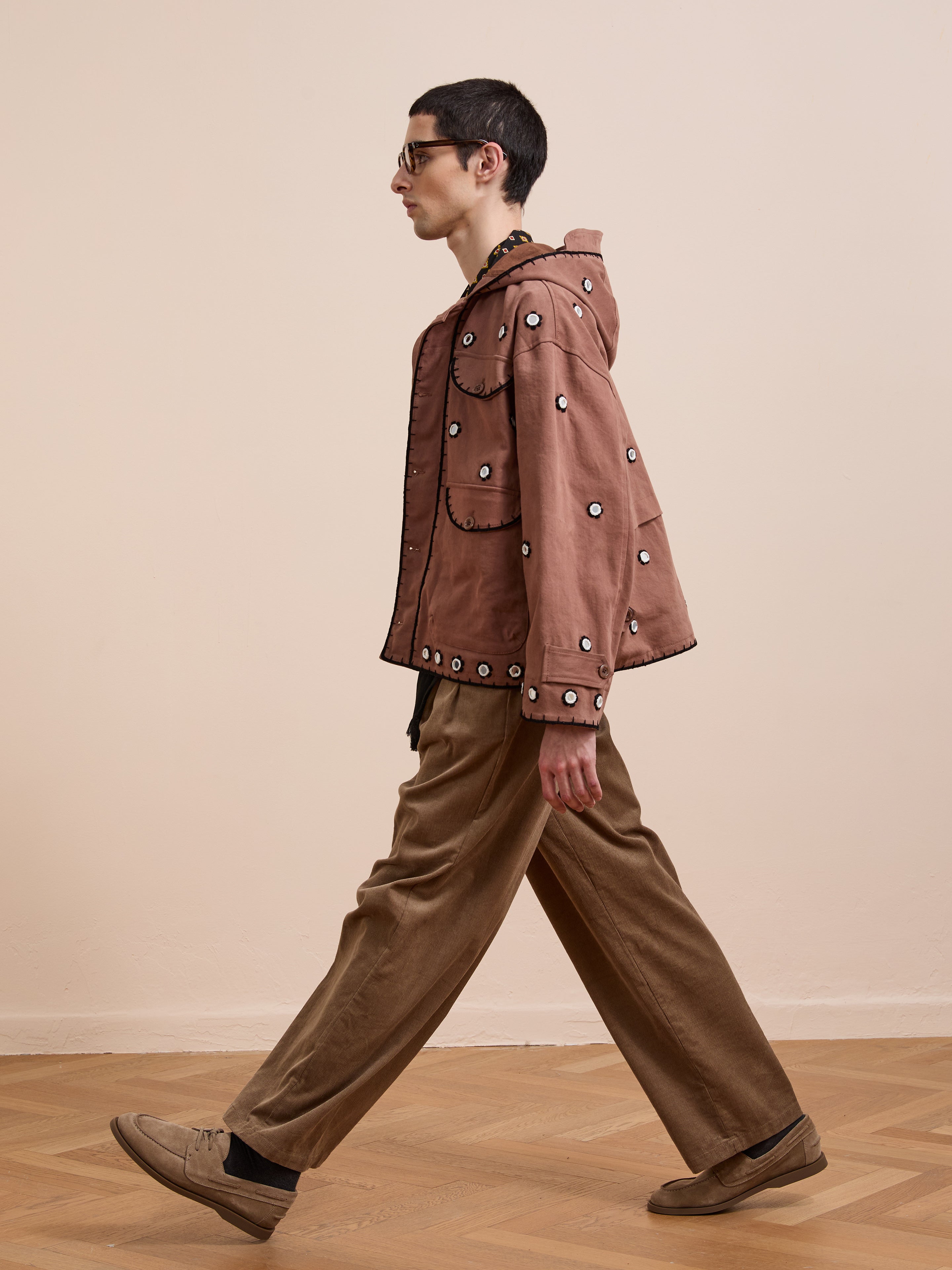 A person with short dark hair and sunglasses walks indoors on a wooden floor against a light wall, wearing the FOUND Hand Embroidered Hooded Mirror Anorak and loose tan pants.