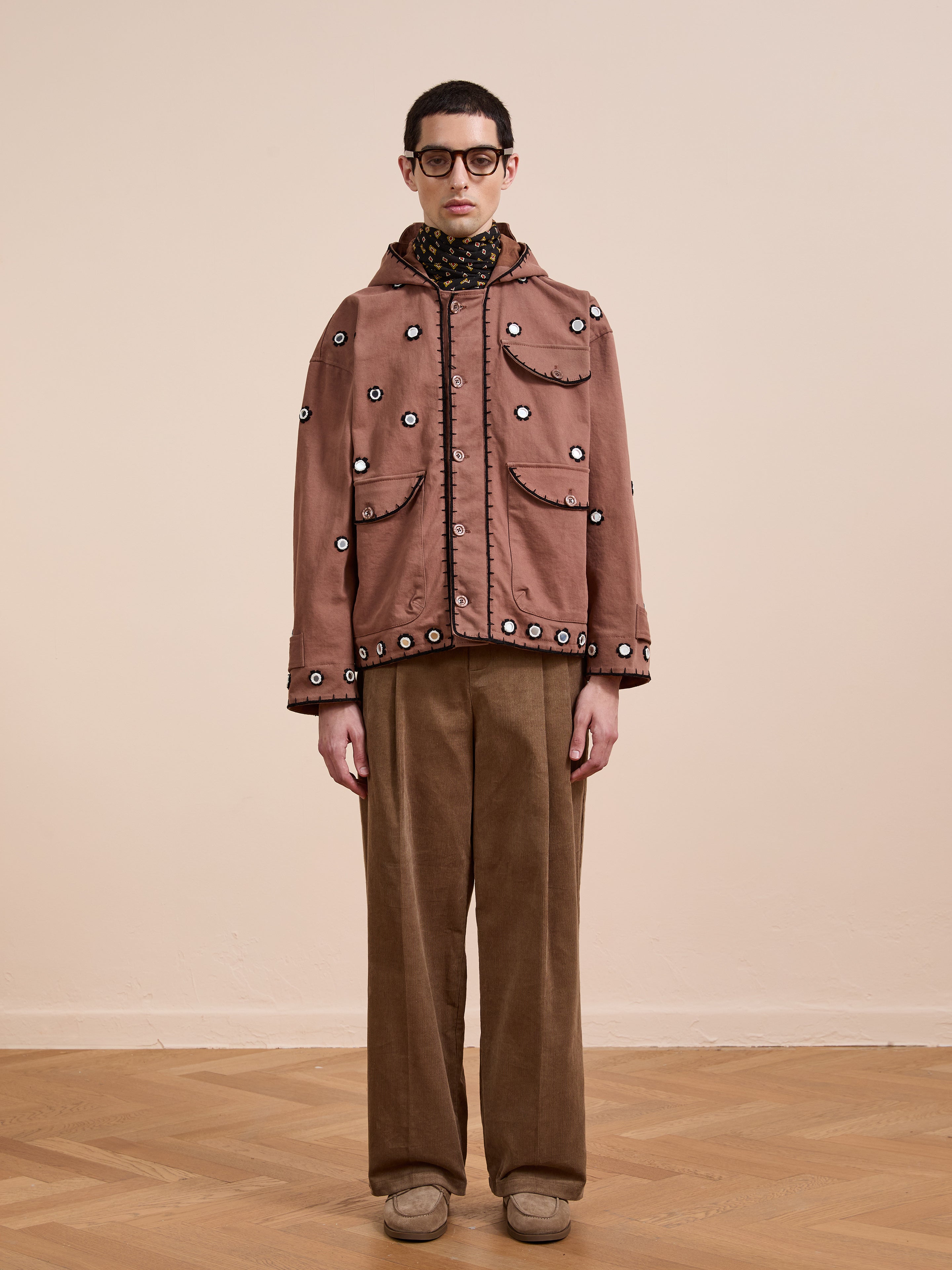 A person stands against a plain background wearing glasses, the FOUND Hand Embroidered Hooded Mirror Anorak, a patterned scarf, brown pants, and beige shoes.