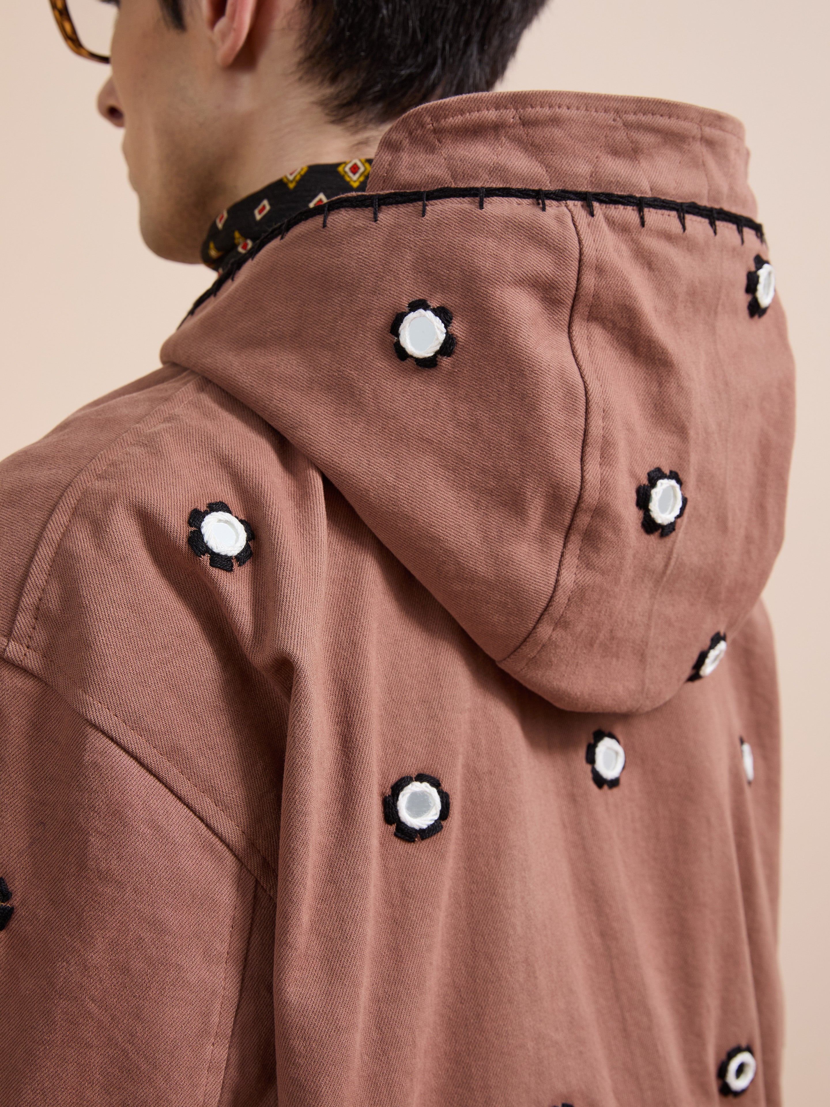 A person is seen from the back wearing the FOUND Hand Embroidered Hooded Mirror Anorak, a brown cotton twill piece featuring black and white circular mirror embroidery.