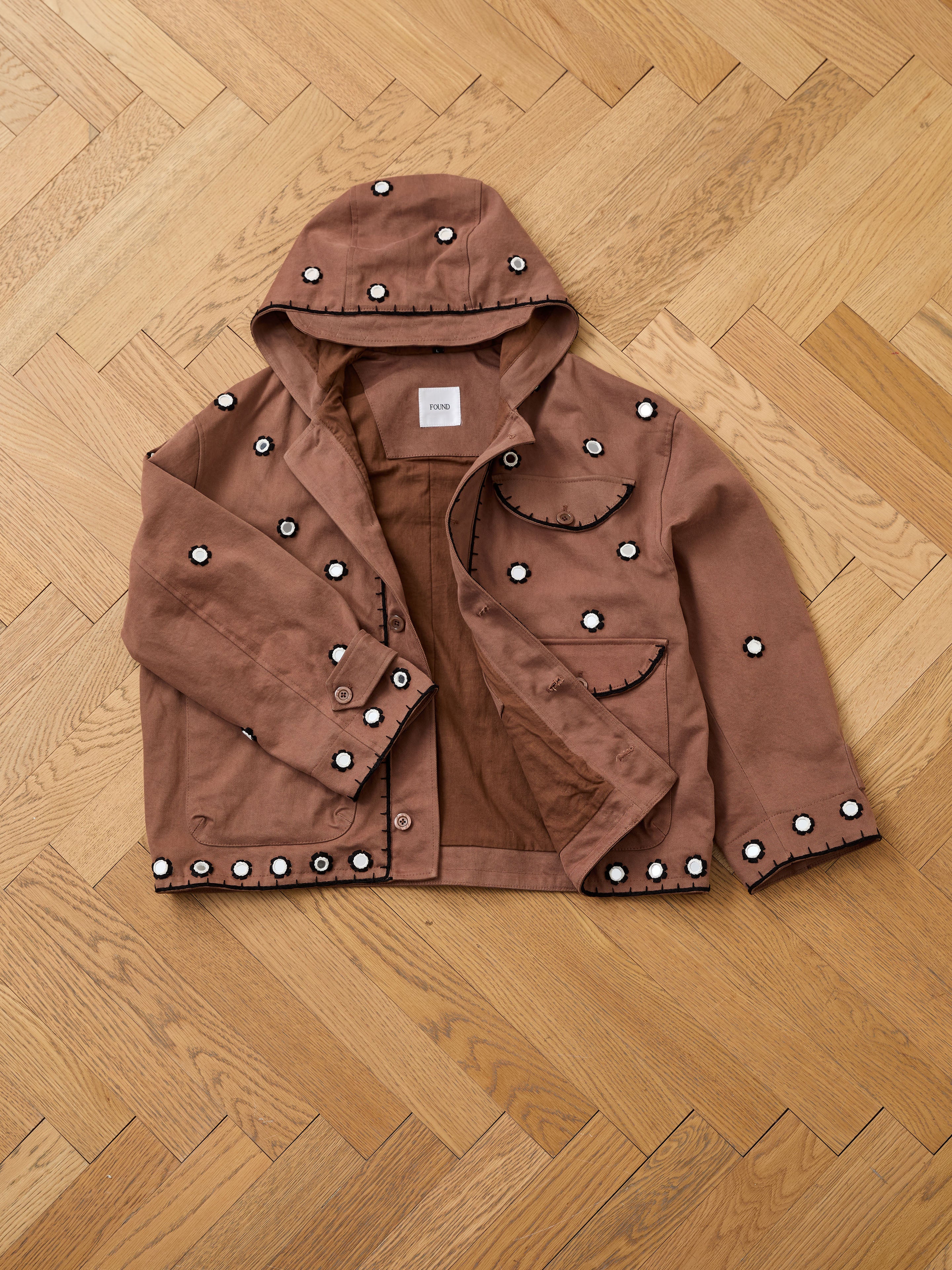 FOUND Hand Embroidered Hooded Mirror Anorak featuring white and black circular designs inspired by South Asian mirror embroidery, shown on a wooden floor.