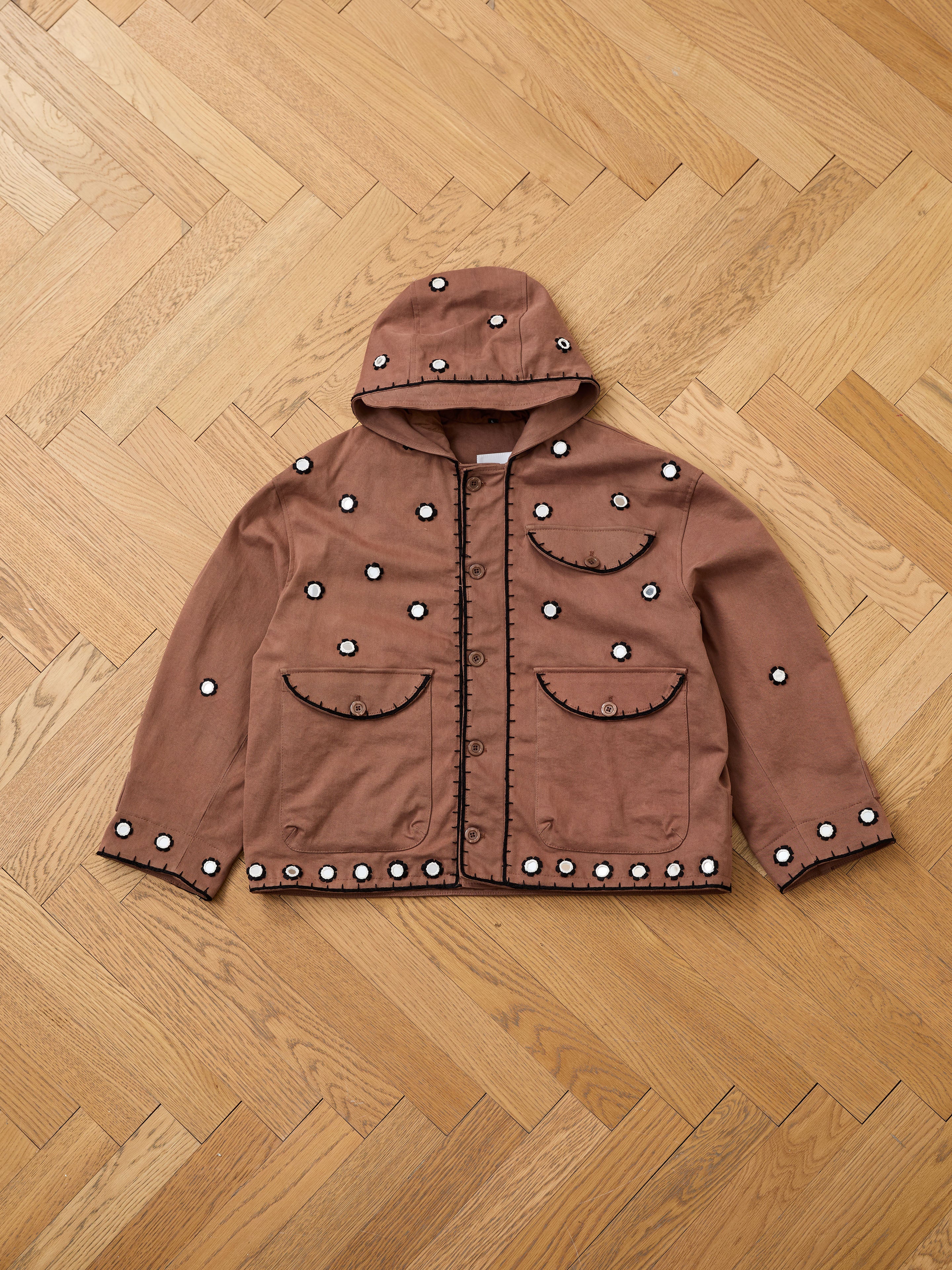 The FOUND Hand Embroidered Hooded Mirror Anorak, in brown with front buttons, two large pockets, and scattered white mirror embroidery, is laid flat on a herringbone wood floor—highlighting South Asian craftsmanship.