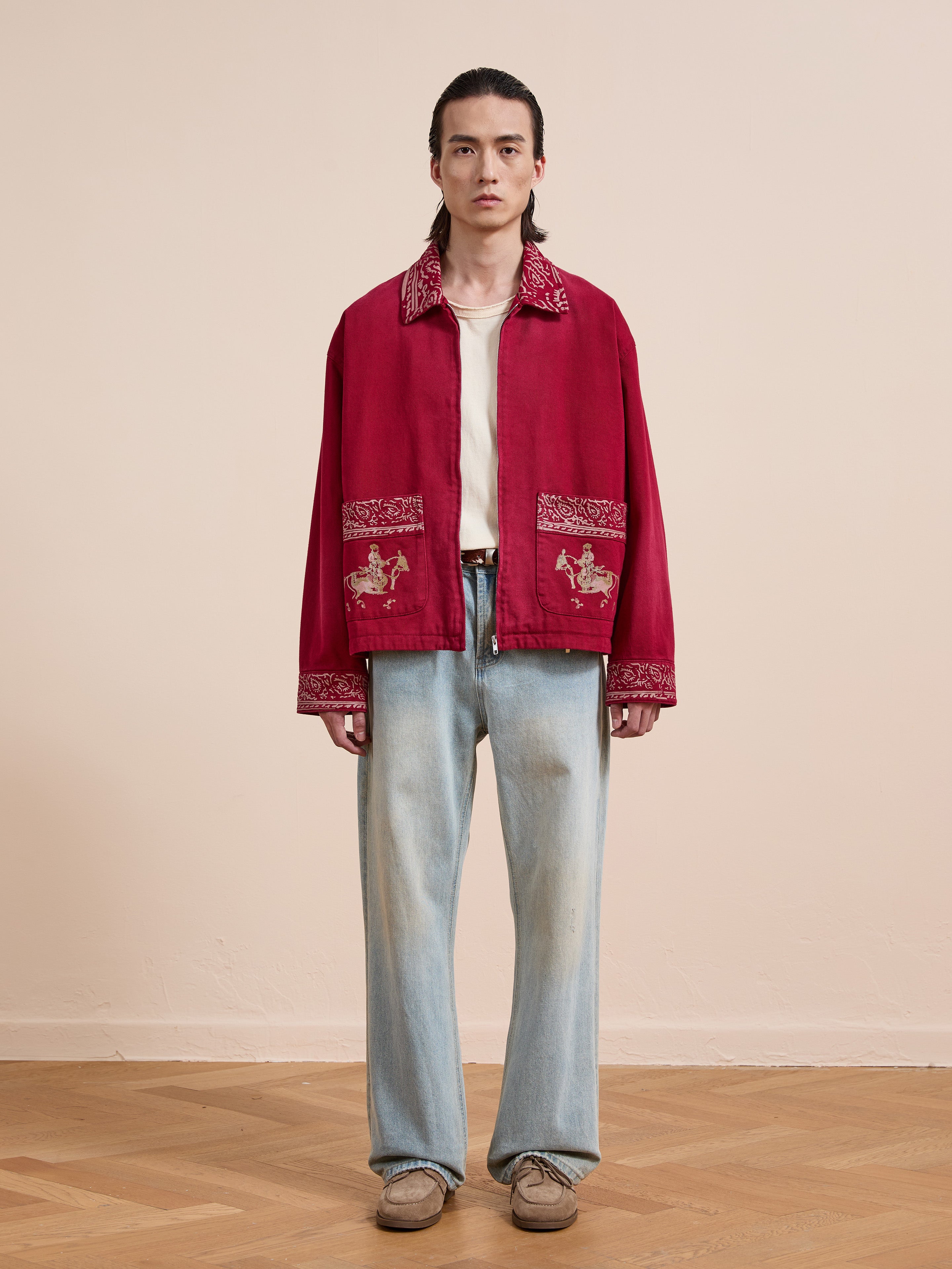 Indoors, a person wears the Found Abhala Printed Work Jacket featuring equestrian motifs, paired with a white shirt, faded blue jeans, and beige shoes against a plain background.