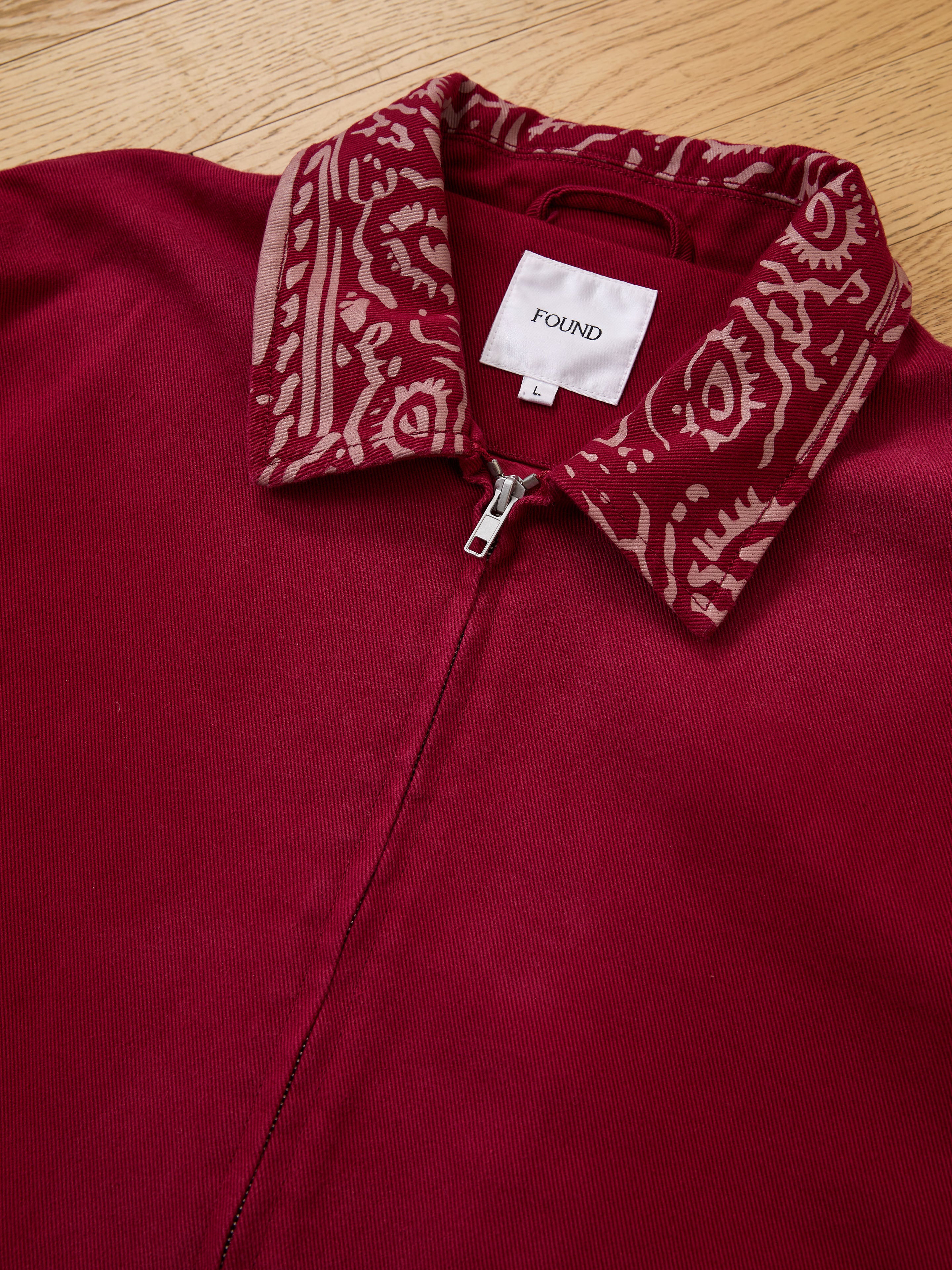 The Abhala Printed Work Jacket by Found, a burgundy zip-up with an equestrian-inspired patterned collar and visible "FOUND L" label, is laid flat on a wooden surface.