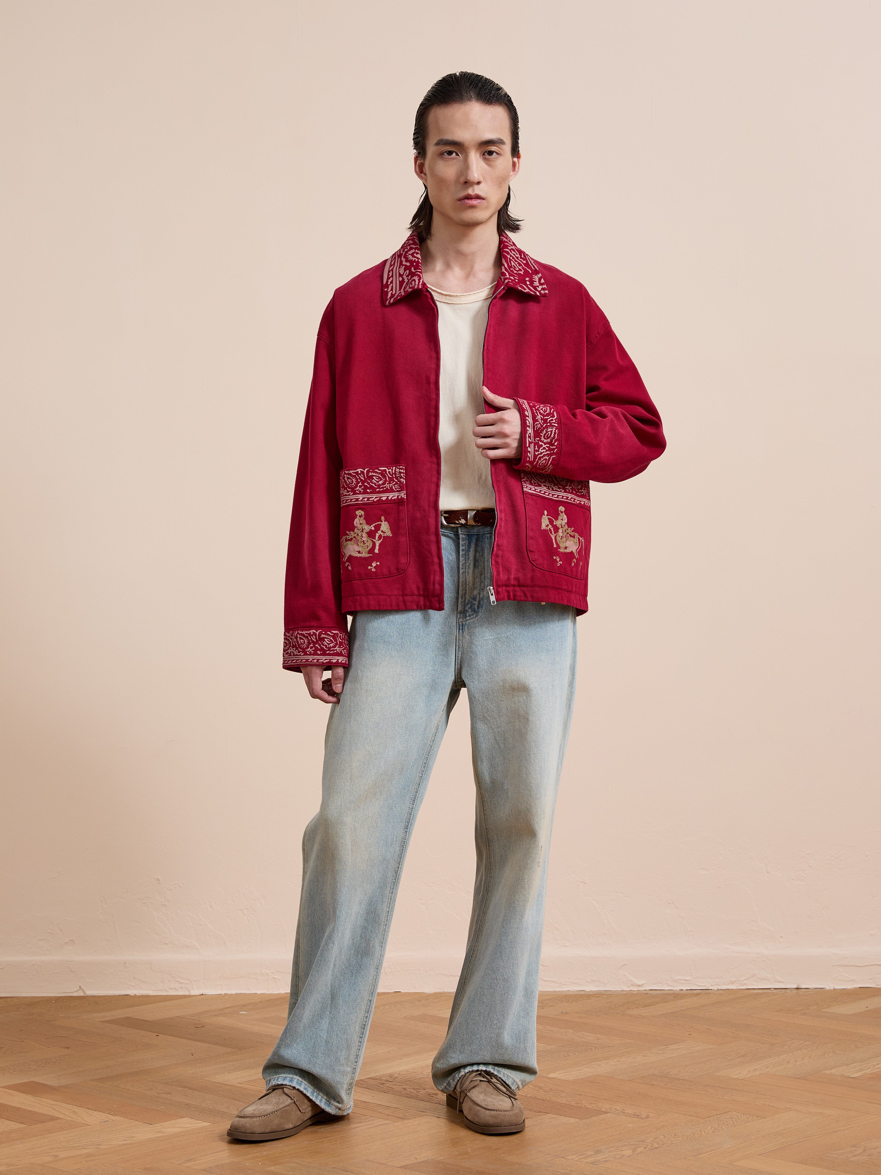 A person stands indoors wearing the Found Abhala Printed Work Jacket, featuring equestrian motifs, paired with a cream shirt, light blue jeans, and brown shoes on a wooden floor against a plain beige wall.